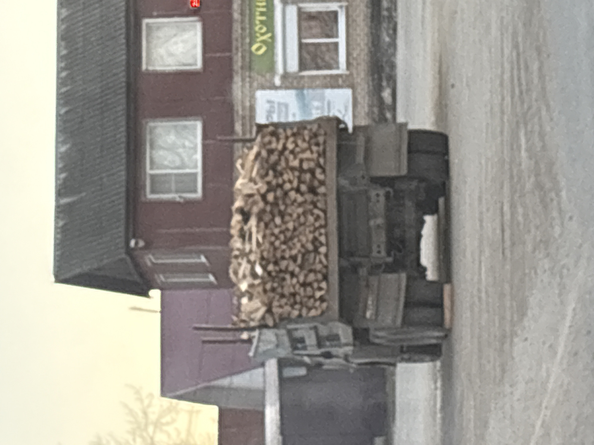 A truck laden with firewood is parked on a street in Chaikovsky, Russia. A building with multiple windows and signs is visible in the background.