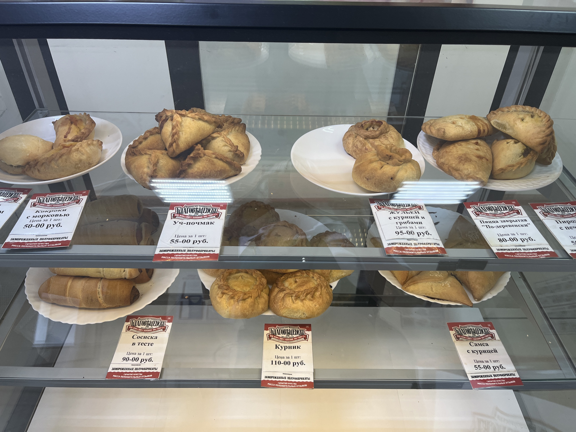 The image shows a display case in a store in Blagoveshchensk, Russia, filled with various frozen pastries. The display case has glass doors and is lit from above. On the top shelf, there are several types of pastries, including triangular-shaped ones filled with what appears to be meat or vegetables, and round pastries with a hole in the center. On the middle shelf, there are several sausage rolls in dough and some round pies. On the bottom shelf, there are more round pies. Each pastry is accompanied by a price tag with Russian text and the store's logo, "БЛАГОВЕЩЕНСК". The overall scene depicts a food retail environment with a focus on baked goods.
