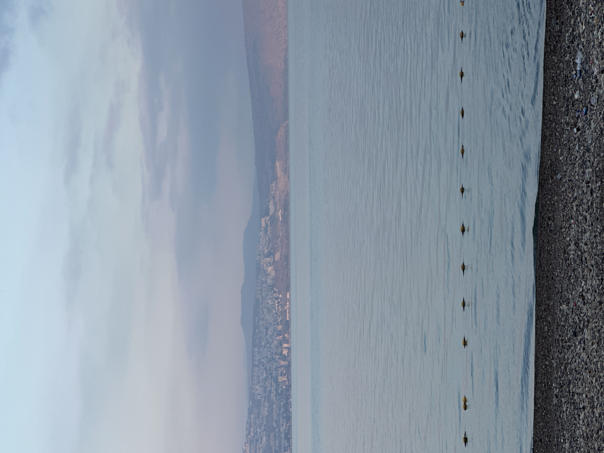 A panoramic view captures the Sea of Galilee, a body of water located in the Emek HaYarden Regional Council, Israel, under a hazy sky. The water's surface is calm, with subtle ripples reflecting the muted light. A line of yellow buoys floats on the water, marking a boundary. In the distance, a city sprawls across the hills on the far shore, its buildings clustered together. Beyond the city, rolling hills form a backdrop, their slopes softened by the atmospheric haze. The foreground features a pebbled beach, with some scattered debris visible near the water's edge. The overall scene evokes a sense of tranquility, with the soft light and muted colors contributing to a peaceful atmosphere.