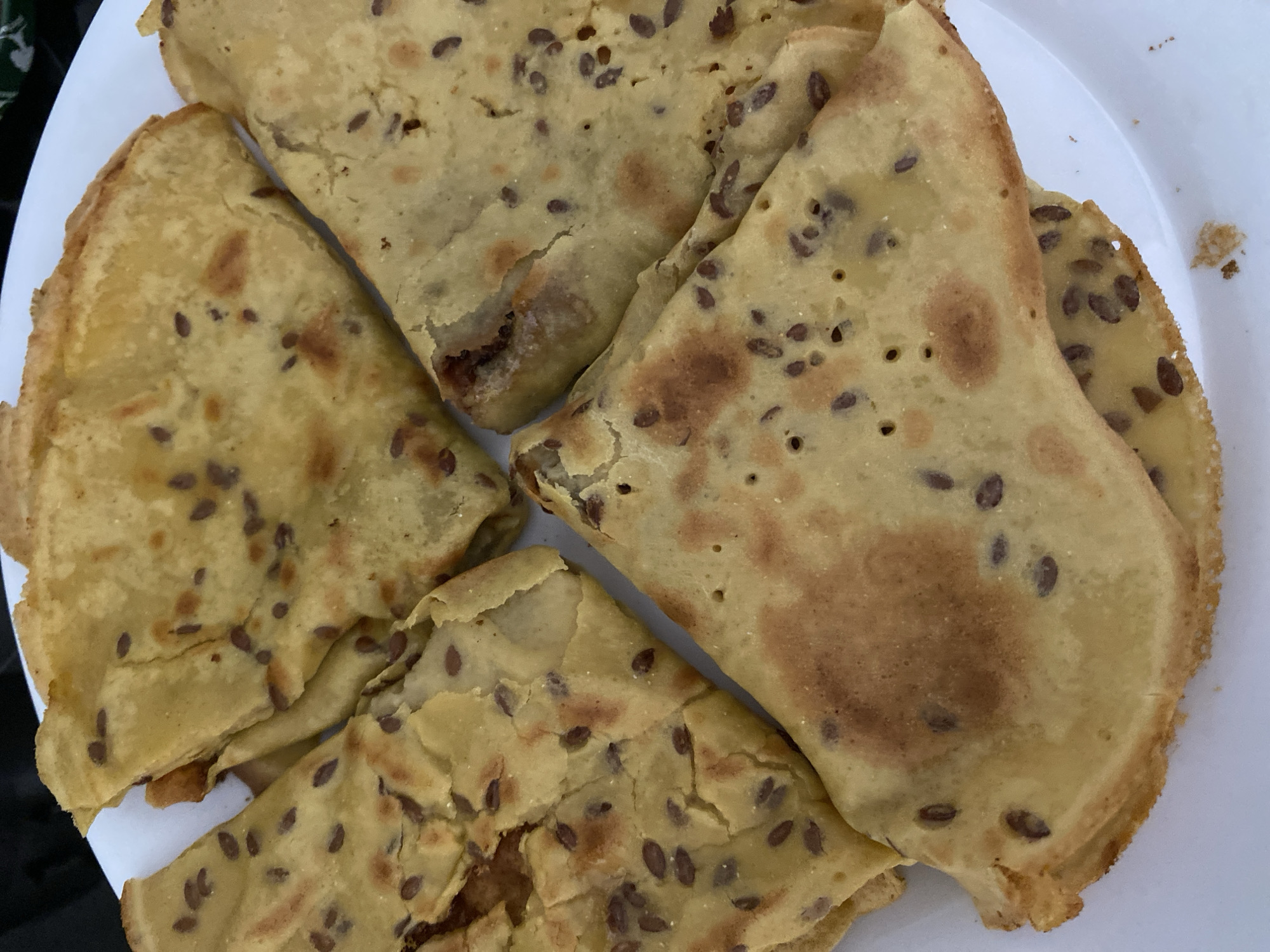 The image is a close-up, overhead shot of several folded crepes or thin pancakes on a white plate. The crepes are light brown with darker flecks of what appear to be flax seeds embedded in the batter. Some areas of the crepes are slightly darker and more browned, indicating they have been cooked. The crepes are folded into triangular or half-moon shapes, overlapping each other. The setting appears to be an indoor environment, possibly a kitchen or dining area, with a dark background visible at the top of the frame. There is no indication of the city of Alanya, Turkey, or any other specific location.