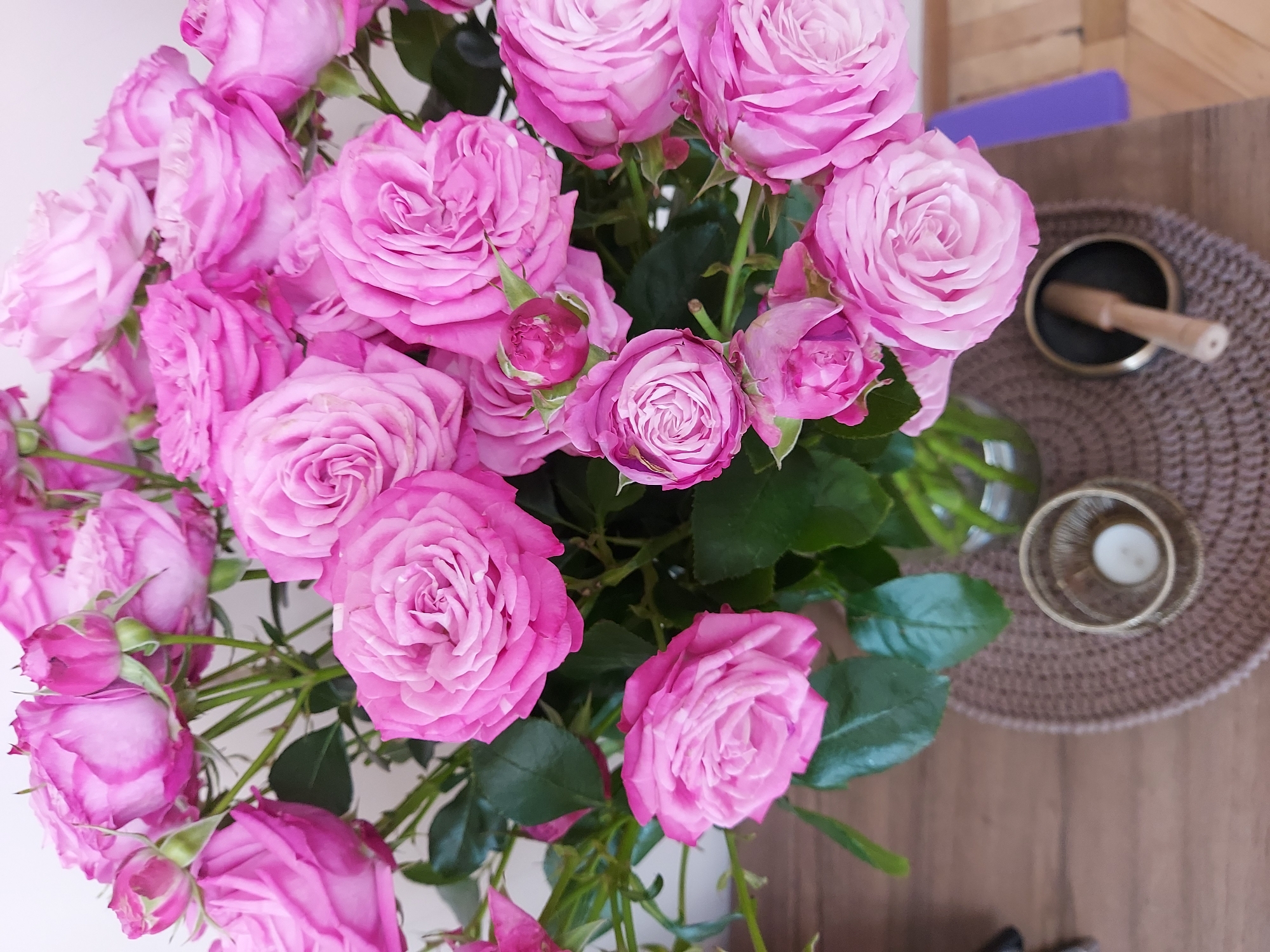 A close-up, high-angle shot captures a bouquet of lush, vibrant pink roses in a glass vase. The roses, with their delicate, layered petals, are the primary subject, filling the frame. The background is softly blurred, suggesting an indoor setting. The arrangement is placed on a wooden surface, with a woven placemat beneath the vase. To the right of the vase, a small, gold candle holder with a white candle is visible, and further to the right, a black bowl with a wooden mallet rests on the placemat. A hint of a purple object is also visible in the far right background. The scene is clean and well-lit, emphasizing the beauty of the flowers. The location is not specified in the image.