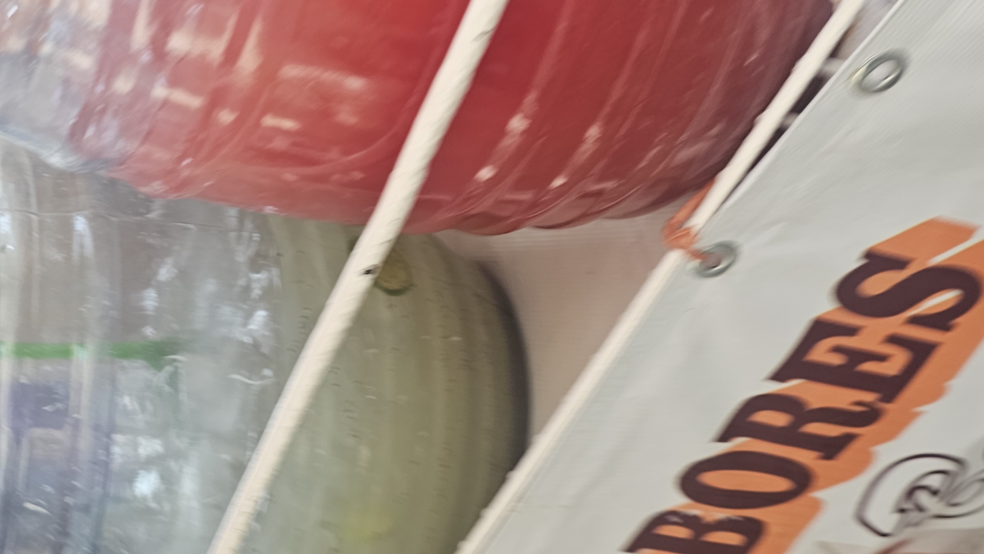 An upward-angled, close-up shot reveals a portion of what appears to be a vendor's stall, likely in a market in Puebla, Mexico. No people are visible in the frame. The scene is dominated by several large containers. At the top, a red plastic container, possibly a jug, is filled with a vibrant red liquid, suggesting a flavored beverage. Below it, a clear plastic container, likely a water jug, is partially visible, filled with a clear liquid. Behind this clear container, an elongated, light green object, possibly a cucumber or a similar vegetable, is present.
To the right, a white banner or sign is prominently displayed, tilted diagonally. It features large, dark red letters with an orange shadow effect. The legible text on the banner reads "BORES". The banner has several metal grommets, through one of which a thin orange string or rope is tied, securing the banner. A thin white stick or pole is visible, running diagonally through the image, separating some of the objects and possibly part of the stall's structure. The lighting indicates it is daytime. The overall impression is that of a refreshing drink stand, possibly selling *aguas frescas* or similar beverages, given the large containers and visible produce.