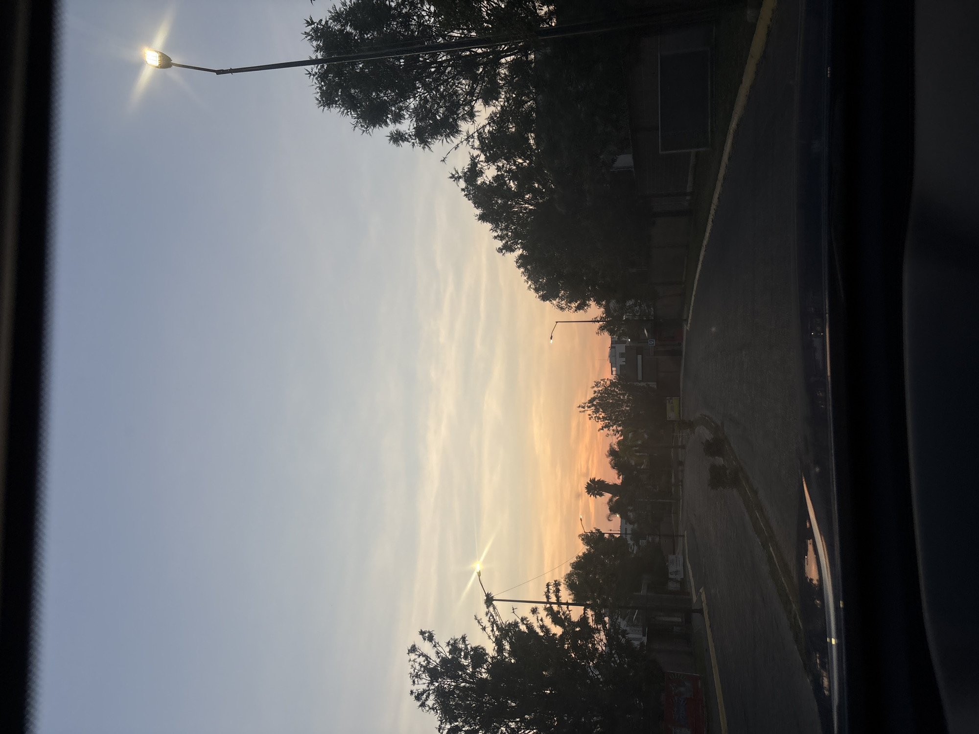 The image depicts a street scene in San Bernardino Tlaxcalancingo, Mexico, at dusk or dawn. The sky shows a gradient of blue to orange, indicating the time of day. Streetlights are illuminated, with one prominently visible in the top left and another near the center of the frame. Silhouetted trees line the sides of the street, adding depth to the view. The road is dark, and a curb separates it from a sidewalk. The photograph appears to have been taken from inside a vehicle, with the car window frame visible on the sides. There are no people evident in the shot.