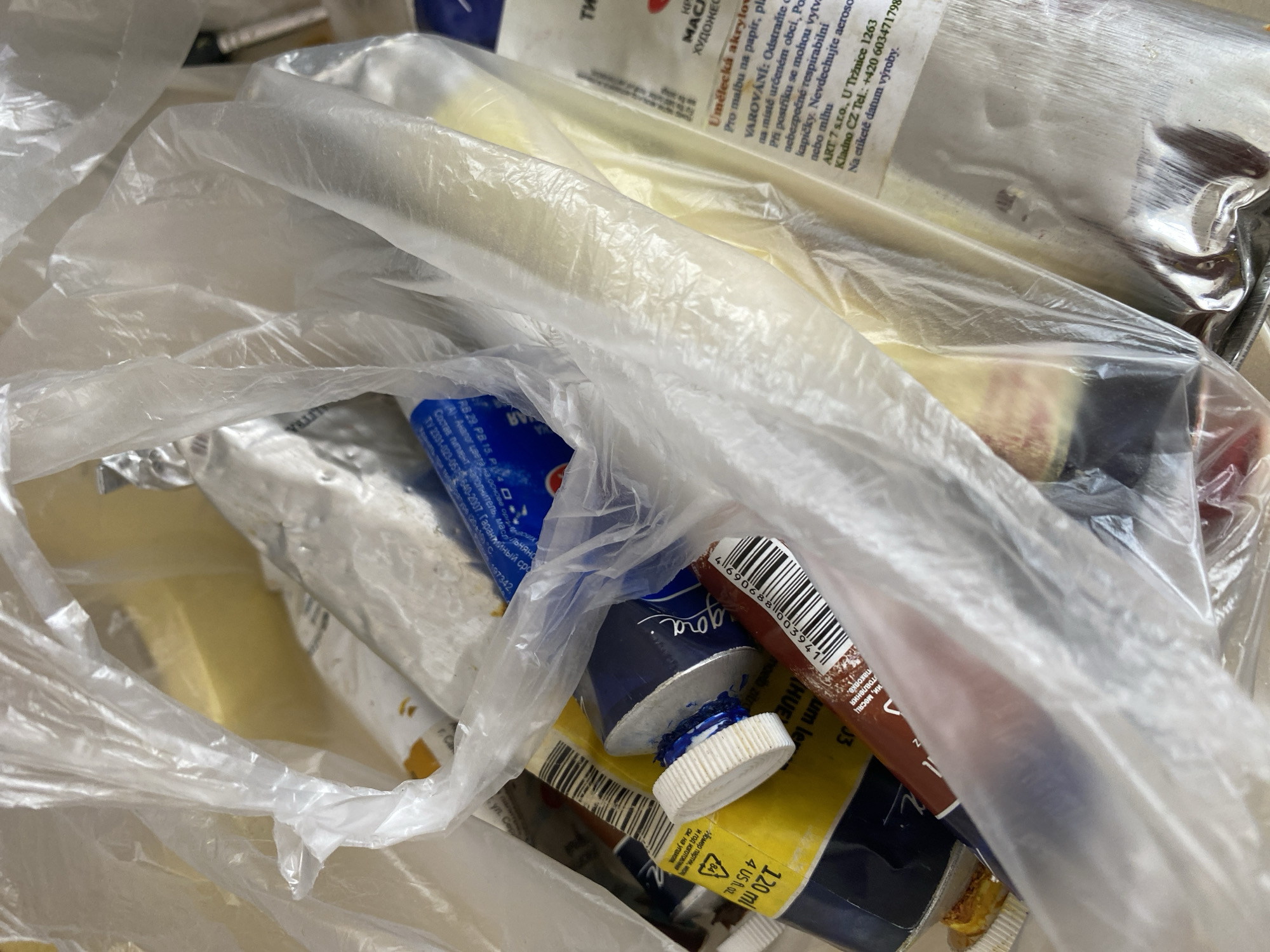 The image shows a close-up, overhead view of several tubes of paint contained within a clear plastic bag. The tubes are of various colors and brands, with some appearing to be oil paints based on the labels and metallic foil packaging. One tube is yellow with "120 ml 4 US FL. OZ." printed on it, and a blue cap. Another tube is dark blue with a silver cap, and some blue paint is squeezed out around the cap. A brown tube is also visible, with a barcode. Some of the tubes have labels with text in what appears to be Czech. A label on one tube reads "Umelecká akrylová" and another section states "VAROVÁNÍ: Odstraňte obal..." which translates to "WARNING: Remove packaging...". The background is out of focus but suggests an indoor setting with neutral lighting. There are no people visible in the image, and no specific location in the city of Bílina, Czechia, can be identified from the visual information provided.