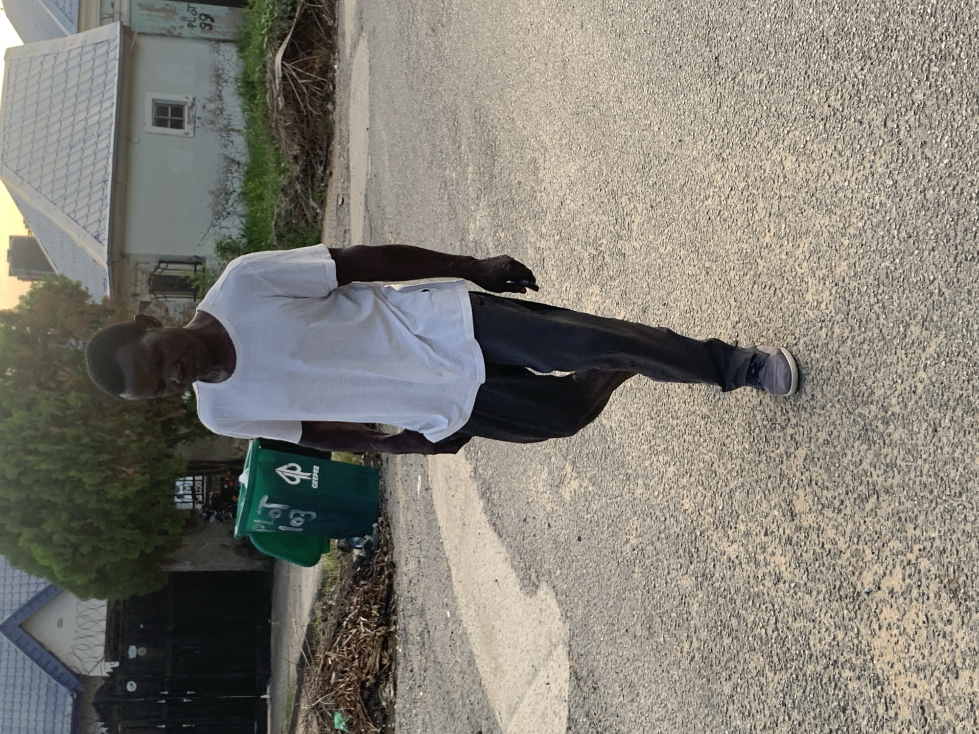 A man walks on an asphalt road. He wears a white t-shirt, dark trousers, and gray sneakers. To the left of the man is a green trash bin with "GEEPEE" printed on it. In the background, there is a house with a gray roof and a window, with "PLOT 99" written on it. There is a black gate with a barbed wire fence behind the bin. The location appears to be a residential street, likely in the city of Abuja, Nigeria, based on the "GEEPEE" trash bin company being located in Nigeria.