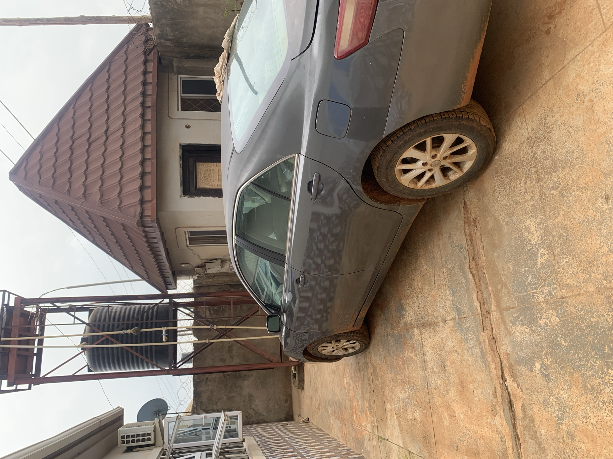 In Kakau, Nigeria, a gray sedan is parked on a dirt-covered concrete driveway in front of a building. The car is viewed from the rear passenger side, showing its right side and rear wheel. The vehicle appears dusty, with dirt splattered on the lower body panels and wheel. A light-colored cloth is draped over the rear windshield.
Behind the car, a tall, dark water tank stands on a metal scaffolding, with pipes extending from it. To the right of the tank is a building with a brown, tiled, pitched roof and a lighter colored facade. A concrete wall, topped with barbed wire, runs along the right side of the property. To the left of the water tank and partially visible is another building with a tiled wall and what appears to be an air conditioning unit mounted on its exterior. The ground is a mix of concrete and reddish-brown dirt. The sky is overcast.