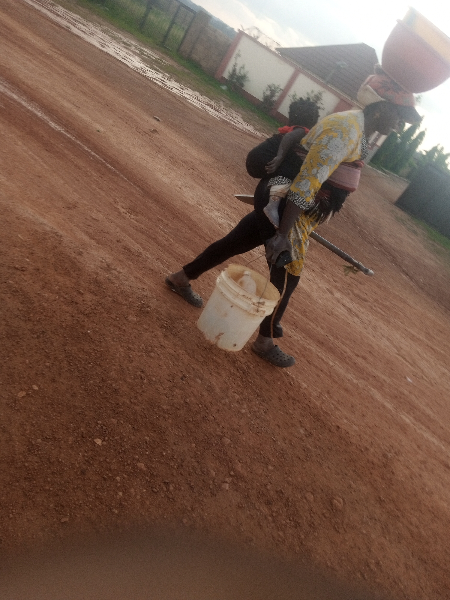 A low-angle, slightly tilted, medium shot captures a person carrying a child on their back and a bucket on the ground. The person, facing right and angled away from the camera, wears a yellow and white patterned blouse and black pants, with gray clogs on their feet. They are carrying a light-colored bucket in their right hand. A child is secured to their back with a cloth wrap. On the person's head, a pink and yellow bowl-shaped object is balanced. The ground is composed of reddish-brown dirt, with patches of water indicating recent rain. In the background, a white wall with a fence is visible, along with trees and a structure. The sky is overcast. The scene appears to be outdoors, on a dirt road or path, likely in a rural or semi-urban setting, potentially in the city of Kwang, Nigeria, given the soil color and vegetation.