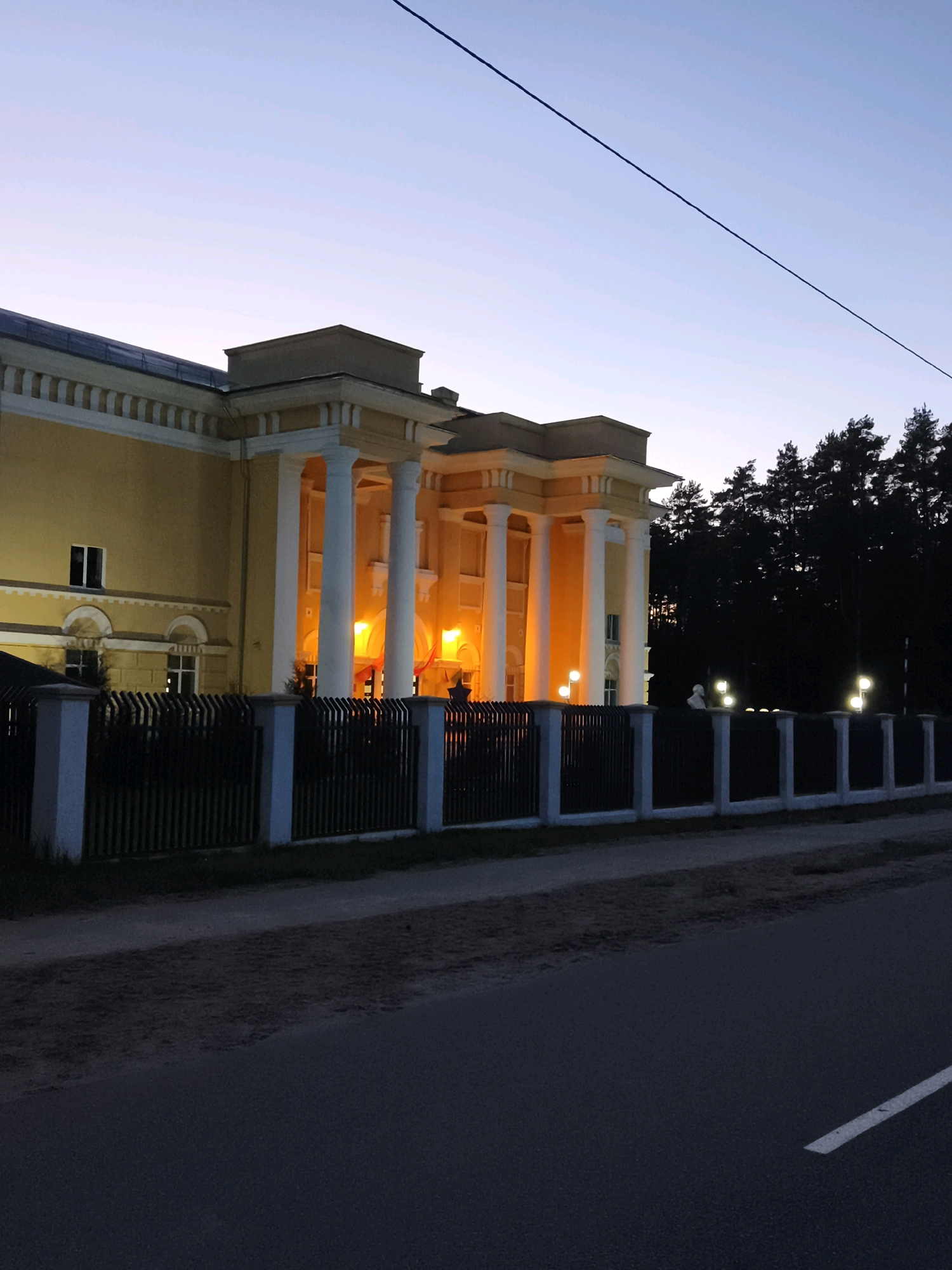 The image shows a stately, yellow, neoclassical building with a colonnade of white pillars illuminated by warm interior lights. The building is set behind a black metal fence with white stone posts. To the right of the building, a dark forest is visible, with several streetlights and a statue of a bust. A power line bisects the pale blue twilight sky. In the foreground, a dark asphalt road runs horizontally across the image, with a white line marking the lane. The scene appears to be a public building in the evening, possibly in the village of Gorski, Belarus, given the prompt. There are no people or animals visible in the frame.