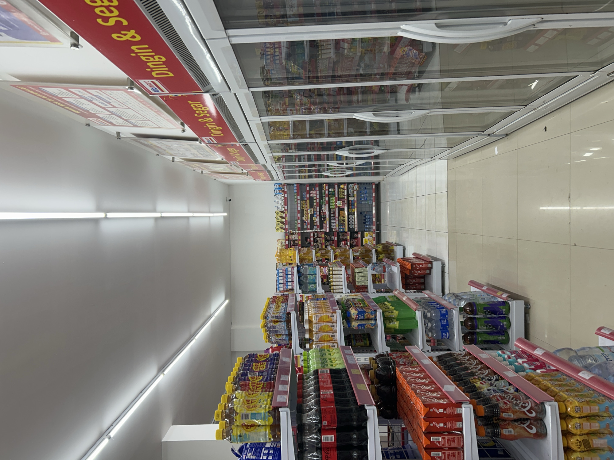 This image captures the brightly lit interior of a convenience store, likely an Alfamart, located in Payadua, Indonesia. The perspective is from an elevated point, looking down a well-stocked aisle.
On the left side of the aisle, rows of shelves display a wide assortment of packaged beverages, including bottled water, various soft drinks, and juices, neatly arranged using gravity-feed systems. Brands like Coca-Cola are discernible. A continuous strip of fluorescent lighting runs horizontally along the upper part of the left wall, illuminating the area. A small security camera is also visible mounted high on this wall.
To the right, a series of large, glass-topped refrigerated display cases extend down the aisle. These coolers house numerous chilled products, with their contents partially visible through the glass, and feature silver handles.
Overhead, red banners with yellow text are suspended from the ceiling, prominently featuring the phrase "Dingin & Segar," which translates to "Cold & Fresh." One banner also clearly displays the "Alfamart" logo, identifying the store chain. Several other white informational signs are also hung from the ceiling.
The floor is composed of light-colored tiles, and the walls are painted in a neutral tone. The store appears clean, well-organized, and efficiently stocked. No people are present in the scene.