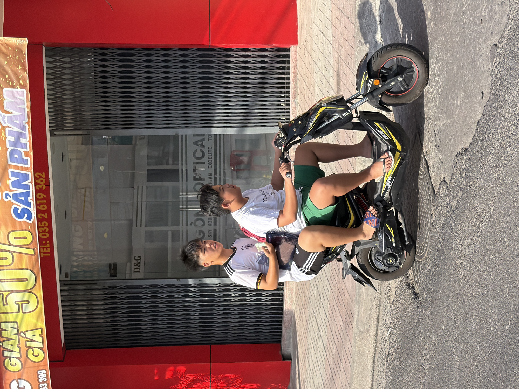 Two young males, appearing to be teenagers, ride a black and yellow electric scooter down a sunlit street in Nha Trang, Vietnam. The rider in front, wearing a white t-shirt with black stripes on the sleeves and black shorts with white stripes, holds a small stack of Vietnamese currency and a brown pouch. The passenger behind him, wearing a white t-shirt and green shorts, appears to be steering. They are positioned in front of a building with a red facade. Above the building entrance, a large banner displays "GIAM 50%" in yellow, "GIÁ" in gold, and "SẢN PHẨM" in blue lettering, suggesting a sale. Below the banner, the phone number "TEL: 035 2 619 362" is visible. The building's entrance features a metal accordion gate, partially open, revealing the interior which has "D&G" and "OPTICAL" signage. The scooter's tires are on a paved surface, with a red and white patterned sidewalk to the right. Shadows from the building and the scooter are cast on the ground.