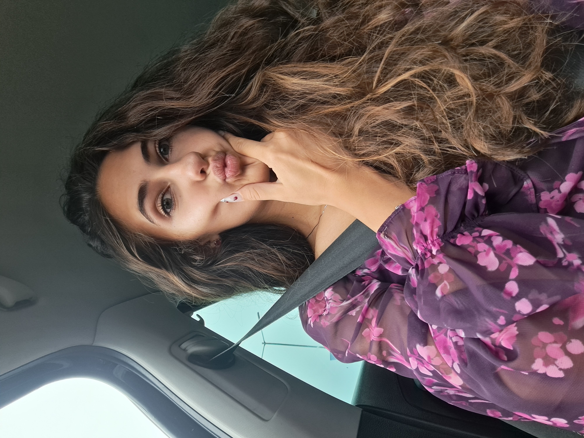 A young woman is captured in a close-up, rotated selfie taken inside a car. Her head is tilted, and her long, wavy hair, a mix of dark brown and lighter highlights, cascades over her shoulder and flows across the frame. She has prominent eyebrows and dark eyes, and her lips are pursed in a "duck face" or "fish kiss" expression. Her left hand is pressed against her left cheek, seemingly to exaggerate the pose, revealing long, manicured nails with white polish and a red dot design on the index finger.
She is wearing a sheer, dark purple blouse adorned with a bright pink floral pattern, resembling cherry blossoms, particularly visible on her ruffled sleeve. A black seatbelt is buckled across her chest, indicating she is a passenger in a vehicle. The car's interior, including part of the headliner and window frame, is visible, with a bright, clear sky visible through the window, suggesting a daytime setting. The woman's expression and pose convey a playful and confident mood. No text is visible in the image.