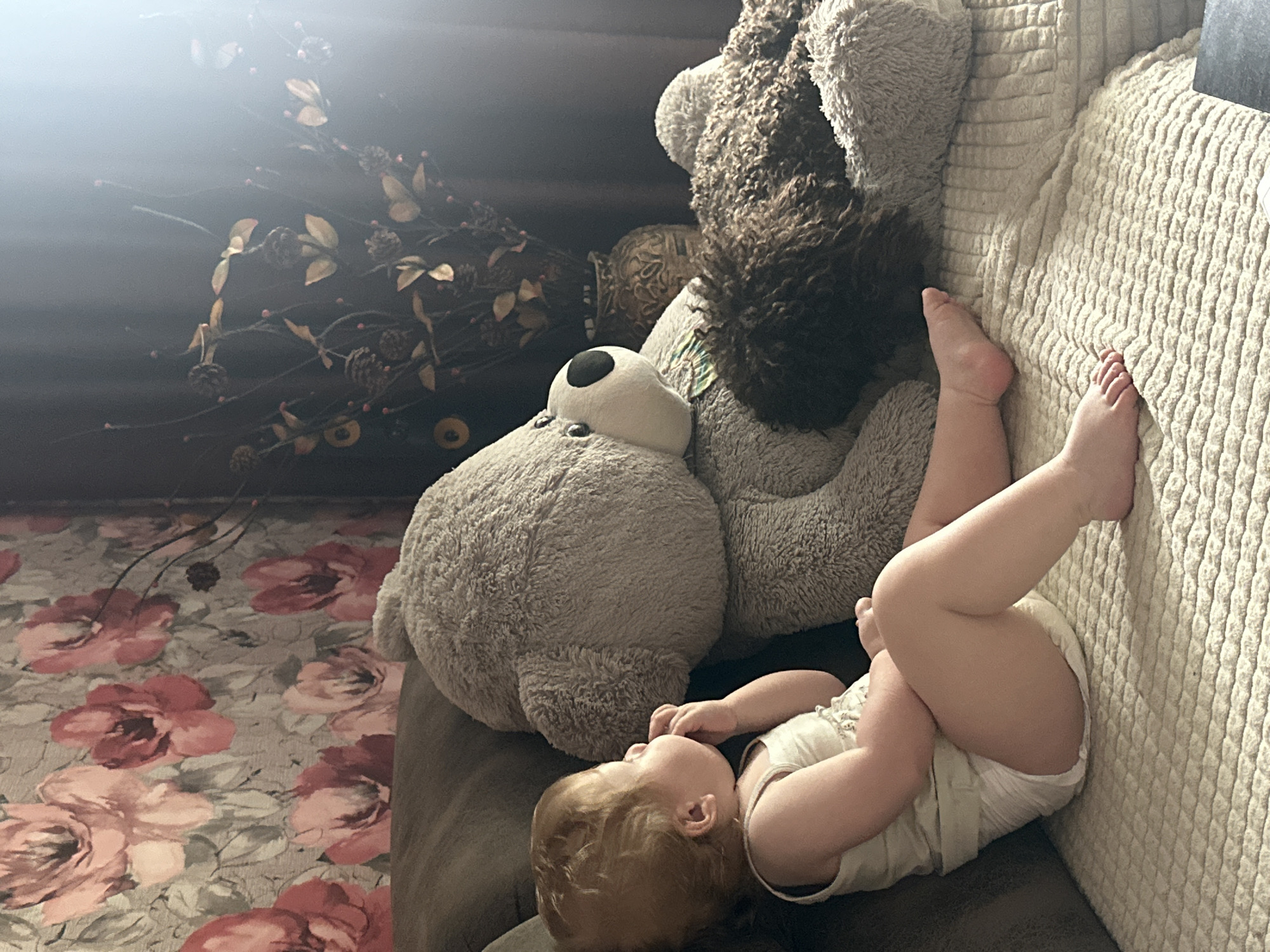 An infant, with blonde hair and wearing a white tank top and diaper, sits on a textured cream-colored blanket. The infant is positioned on the left side of a sofa, with their back to the camera and head turned to the right, appearing to suck on their fingers. Adjacent to the infant is a large, light gray stuffed bear. To the right of the large bear, and partially behind it, is a smaller stuffed dog with dark, curly fur. The background features floral wallpaper with pink and red flowers, a vase with dried floral arrangements and pinecones, and a window with bright light streaming in from the right. The scene is likely indoors, in a residential setting within the city of Sayanogorsk, Russia.