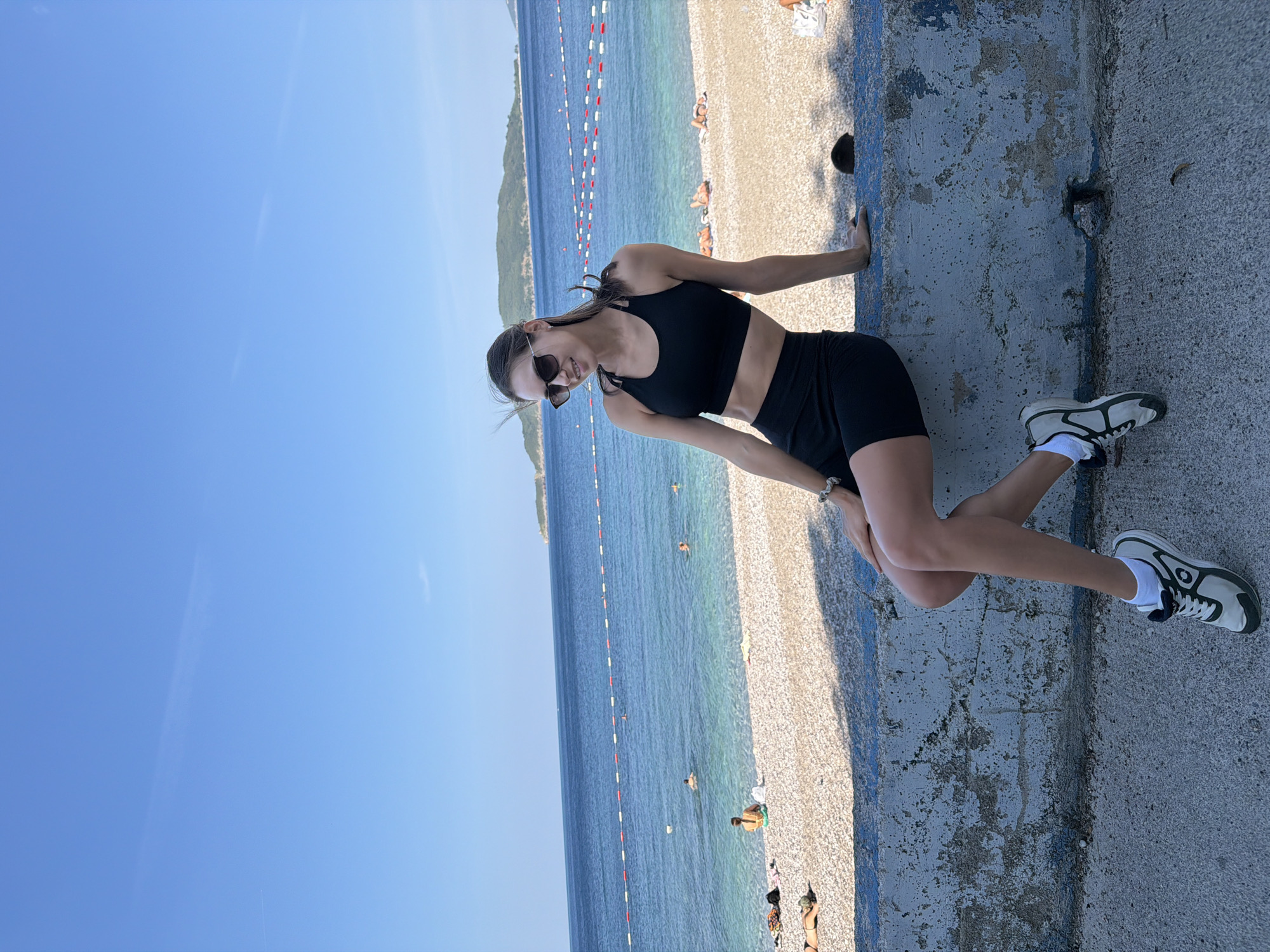 A young woman is seated on a concrete wall overlooking a beach in Kamenovo, Montenegro. The woman is wearing a black sports bra and black biker shorts, with sunglasses and athletic shoes. The beach has a pebble shore, with several people sunbathing and swimming in the clear blue water. The sea stretches to the horizon, meeting a clear blue sky. In the background, a distant island is visible. The sun is shining brightly, casting shadows on the scene.