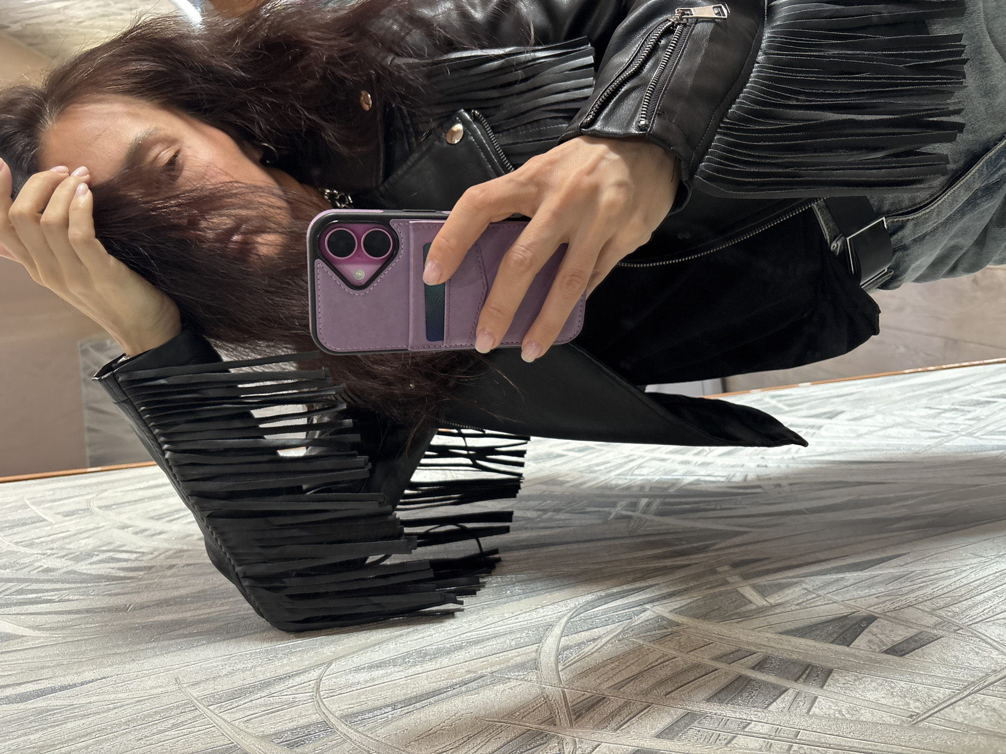 An individual, likely female, with long, dark hair is captured in what appears to be a mirror selfie. Her head is tilted, with hair partially obscuring her face, and her eyes are closed or cast downwards. She is wearing a black leather jacket featuring prominent fringes on the sleeves and shoulder area, along with visible zippers and buttons. A pair of jeans can be seen on her lower half.
In her right hand, she holds a smartphone encased in a distinctive purple wallet-style phone case. The case has compartments, and what appears to be a card is visible in one slot. The phone's camera lenses are also visible through the case. The background suggests an indoor setting with a patterned surface, possibly a wall or floor, reflecting the person's image. The overall orientation of the image is rotated, common in mirror selfies. No specific text is visible in the image. The location is Chaikovsky, Russia.