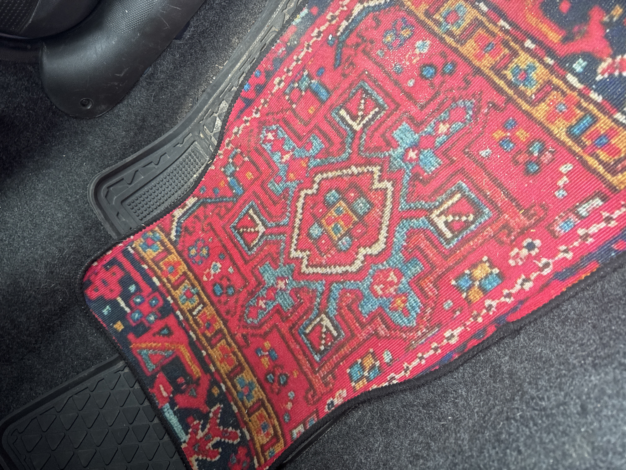 A red oriental rug with a black border is placed on a dark gray car carpet in the footwell of a vehicle. The rug has an intricate geometric pattern in blue, yellow, and white. To the left of the rug is a black rubber car mat with a diamond-shaped tread pattern. To the right of the rug is a black plastic car interior component, possibly part of the center console or seat base. The scene is indoors, within a vehicle, and does not provide any specific details about the city of Sofia, Bulgaria.