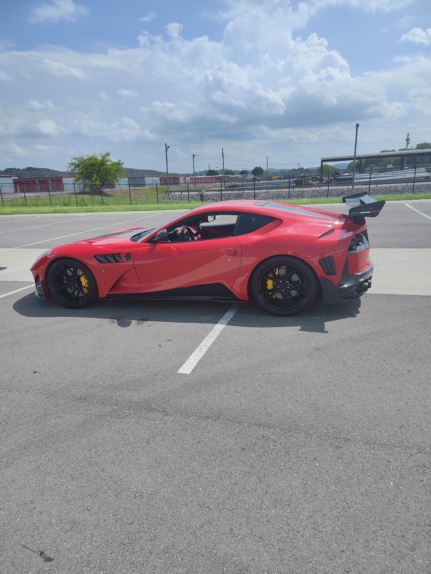 A bright red, highly modified sports car is parked in an asphalt parking lot. The car has a prominent rear spoiler with "STALLONE" written on it, black wheels with red accents, and yellow brake calipers. The car's body features aggressive aerodynamic enhancements, including large air vents and a splitter. The parking lot has white lane markings and appears to be outdoors. In the background, a chain-link fence separates the parking lot from a grassy area with storage units and some trees. Beyond that, there are power lines and a gently rolling landscape under a partly cloudy sky. The image seems to have been taken in a location that could be the city of Lebanon, United States of America, given the overall aesthetic of the surroundings. There are no people visible in the image.