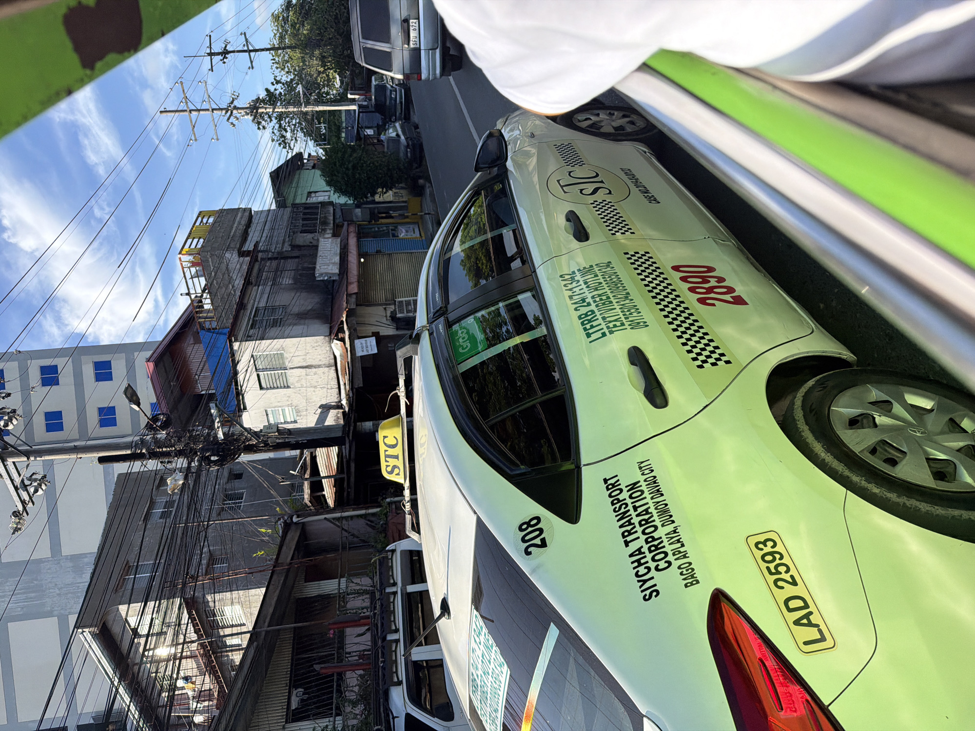 In Davao City, Philippines, a light green taxi with the number 208 and license plate LAD 2593 is visible on a street. The taxi has "SIYCHA TRANSPORT CORPORATION BAGO APLAYA, DUMOY DAVAO CITY" written on its side. A yellow sign on the taxi roof displays "STC". Other vehicles, including a silver van with license plate SFU 672, are also present on the road. The background features multi-story buildings, power lines, and a clear blue sky with some clouds. A person's white shirt sleeve is partially visible in the foreground, suggesting the photo was taken from another vehicle.
