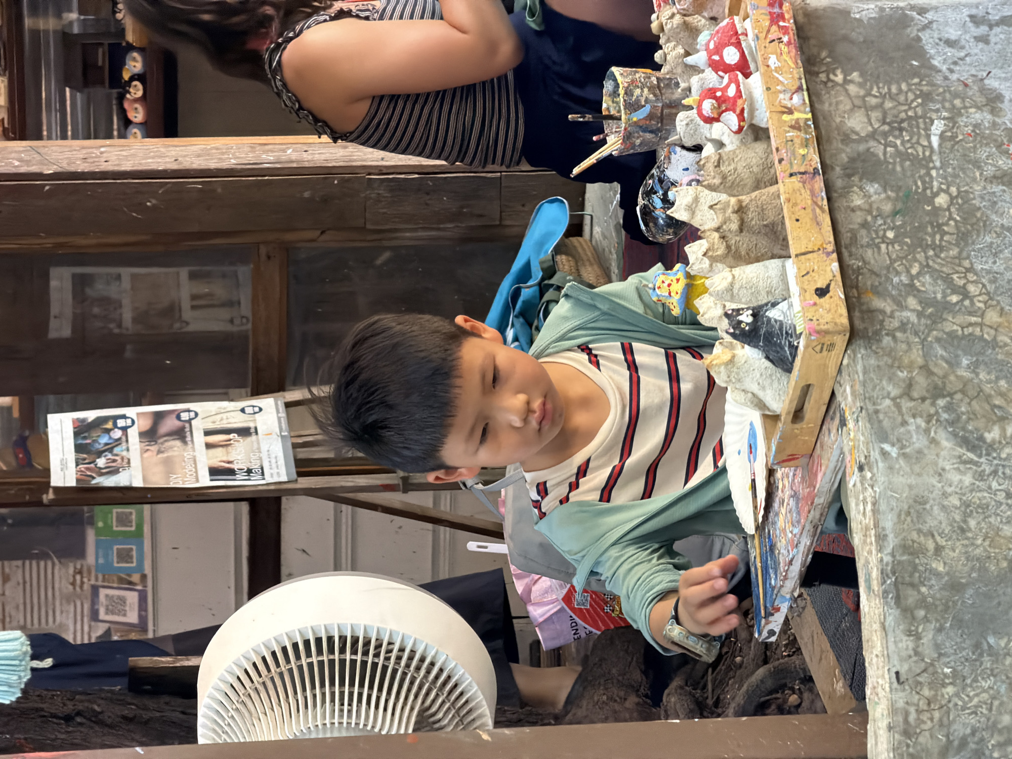 This image shows a young East Asian boy sitting at a table, engaged in a craft activity. He is wearing a striped t-shirt and a light green jacket, and a watch on his left wrist. In front of him is a tray containing several small, unpainted or partially painted figures, including cats, mushrooms, and other abstract shapes. There are also art supplies, such as brushes and a cup with paint, on the table.
Behind the boy, a woman with dark hair is visible, seated and also seemingly involved in a craft. The background reveals a rustic wooden structure, possibly an outdoor market stall or workshop, with various items displayed on shelves. The presence of QR codes on a nearby display suggests this location is in Thailand. The scene is located in Ban Sai Kham, Thailand, a village known for its crafts and cultural tourism. The overall atmosphere suggests a creative and engaging environment for artistic pursuits.