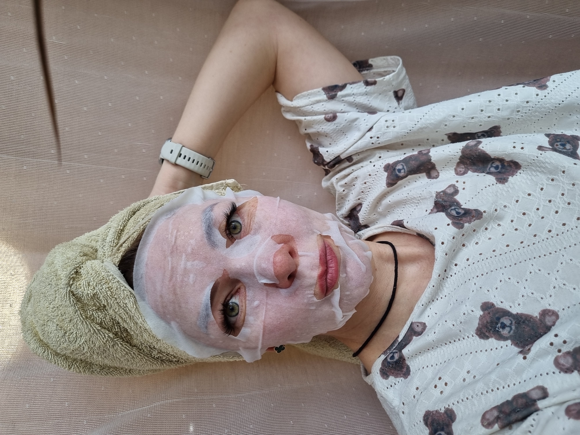 A woman lies on a light-brown textured surface, possibly a bed or couch. She is wearing a white, short-sleeved top with a pattern of brown teddy bears and a black necklace. She has a white sheet mask on her face and a light-green towel wrapped around her head. Her left arm is raised, showing a light-colored smartwatch on her wrist. The location is Chaikovsky, Russia.