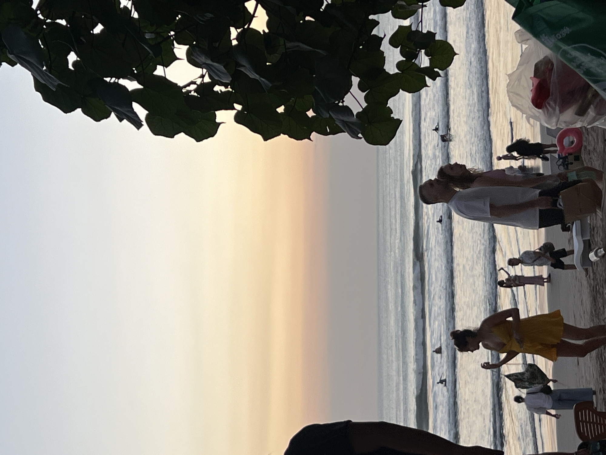 The image depicts a beach scene in Kuta, Indonesia, during what appears to be sunset. A tree with large, dark green leaves dominates the right side of the frame, obscuring a portion of the sky and the horizon. The sky transitions from a pale blue at the top to soft hues of pink and orange closer to the horizon. The ocean is visible with gentle waves rolling in, and several surfers are scattered in the water, some appearing to be standing on their boards.
On the sandy beach in the foreground, a woman in a bright yellow dress is the central figure, looking to her left with her hand slightly raised. Behind her, a couple walks side-by-side, the man in a white t-shirt and the woman in a pink top. Further back, other beachgoers are visible, including individuals walking and some silhouetted figures. A man in a light-colored shirt and shorts walks towards the right, carrying a bag. Towards the far left, the shoulder and arm of a person in a black shirt are partially visible. In the foreground, a plastic chair is partially visible on the lower left, and to the right, several plastic bags and what appears to be a pink inflatable ring are scattered on the sand. The overall atmosphere suggests a relaxed evening at the beach.