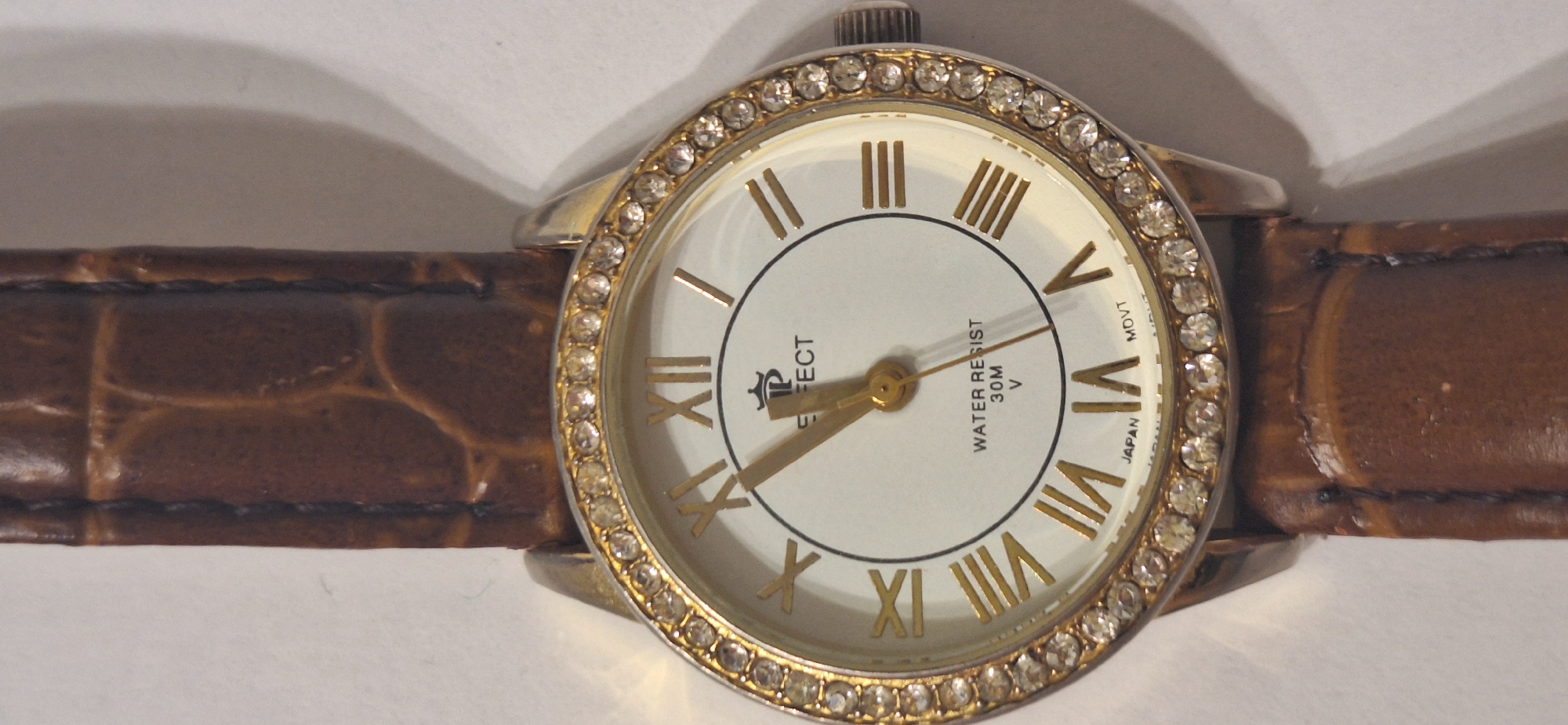 A close-up shot focuses on a woman's wristwatch. The watch features a white face with gold Roman numerals and hands. A decorative band of clear rhinestones encircles the face, and a brown, textured leather strap is attached. The watch is set against a plain white background. No city location is visible.