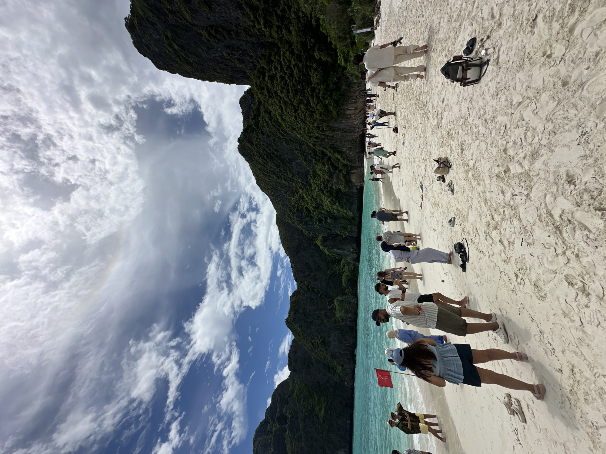 The image captures a bustling beach scene at Maya Bay, located within the Phi Phi Islands, near Ao Nang, Thailand. The foreground is dominated by a white sandy beach with a scattering of flip-flops and bags. Several groups of people are present, mostly tourists, dressed in casual beachwear and some in more formal attire, suggesting a range of activities. Some are walking along the shore, while others are standing in small groups, conversing or observing the surroundings. The turquoise waters of the Andaman Sea gently lap the shore, and a red flag is planted on the beach, possibly indicating a warning or a designated area. Towering limestone karsts covered in lush green vegetation form a dramatic backdrop to the bay, characteristic of the Phra Nang Cave Beach area. The sky is a mix of bright blue and scattered white clouds, with a subtle rainbow visible in the upper left quadrant. The overall atmosphere is one of a popular tourist destination experiencing a day with varied weather conditions.