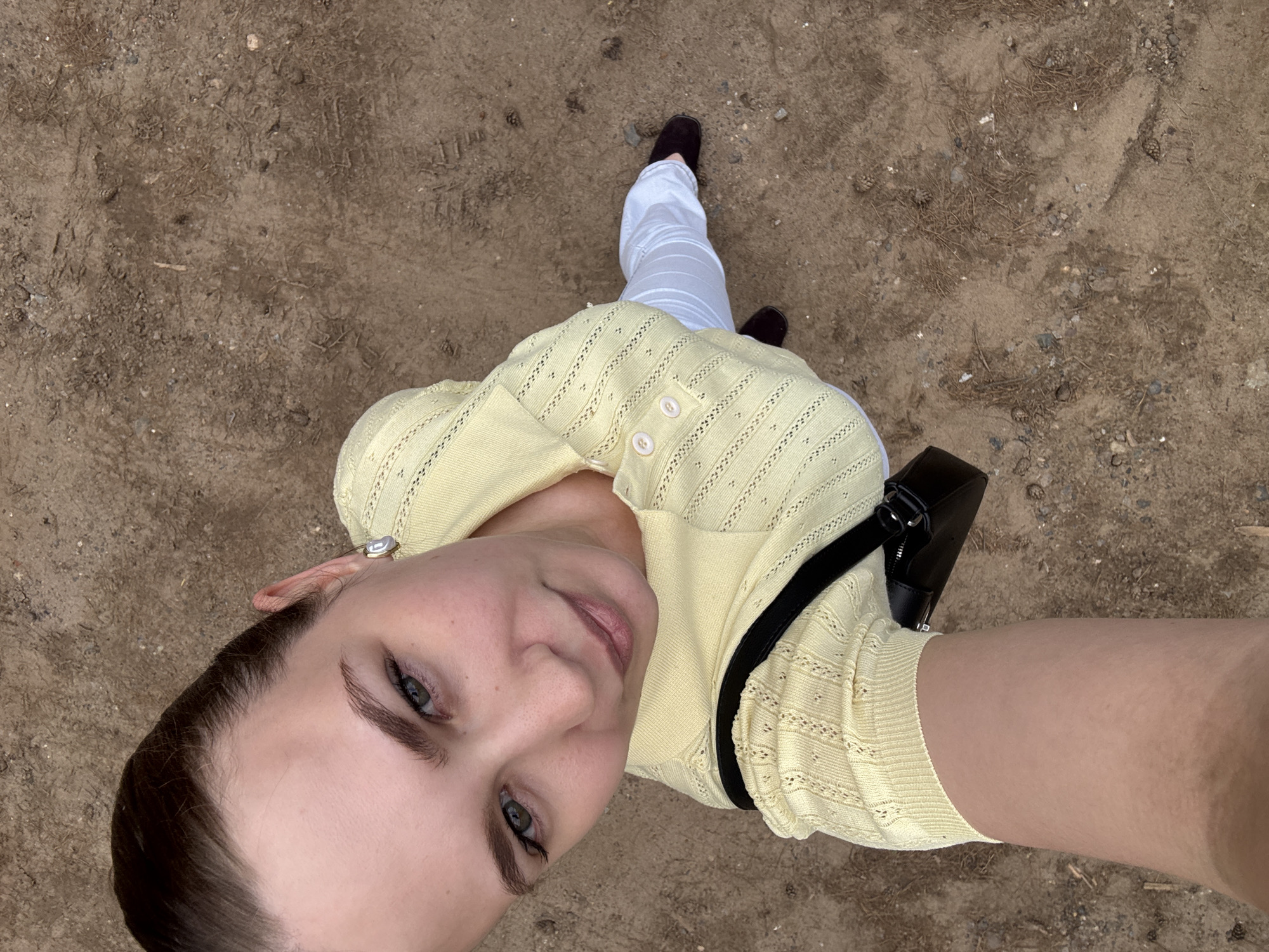 An overhead selfie captures a young adult female standing on an unpaved ground. She has light skin, dark hair pulled back from her face, and greenish-blue eyes that look directly at the camera. She wears a pale yellow short-sleeved knit polo shirt with horizontal openwork patterns, a small collar, and three white buttons down the front placket. Below, she wears white trousers and dark brown flat shoes. A black shoulder bag with a visible strap is draped over her left shoulder. The ground is composed of reddish-brown soil, mixed with small gravel, pebbles, dry pine needles, and several small pine cones. There are no visual elements within the image to confirm or deny the location being the city of Chaikovsky, Russia.