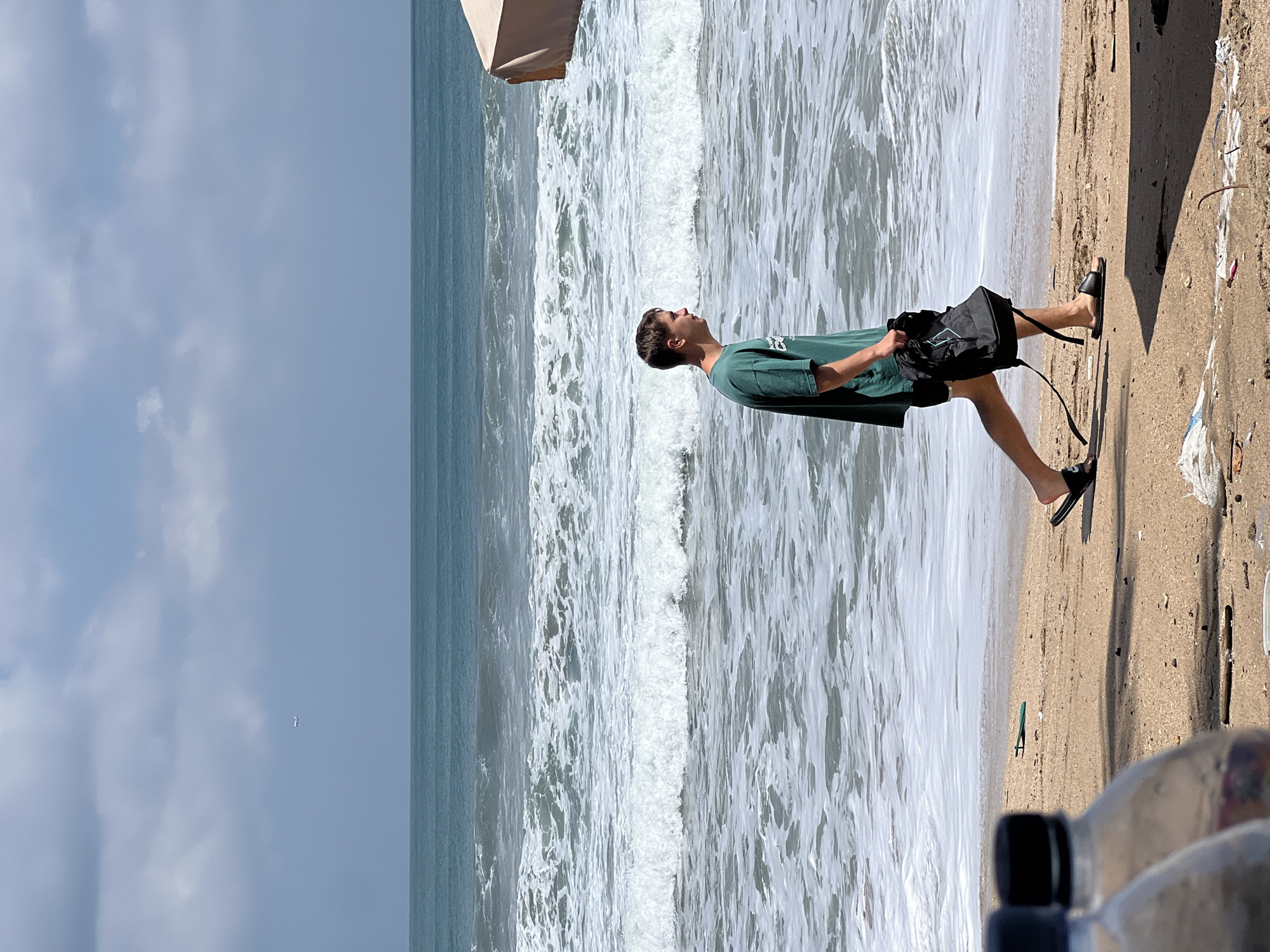 A young man walks along the shore in Kuta, Indonesia, carrying a black backpack. He is wearing a green t-shirt and black shorts, with black sandals on his feet. The ocean waves crash onto the sandy beach, and the sky is cloudy. In the distance, a small airplane is visible in the sky. The scene depicts a typical day at the beach, with the man enjoying a stroll by the sea. The overall impression is one of relaxation and natural beauty.
