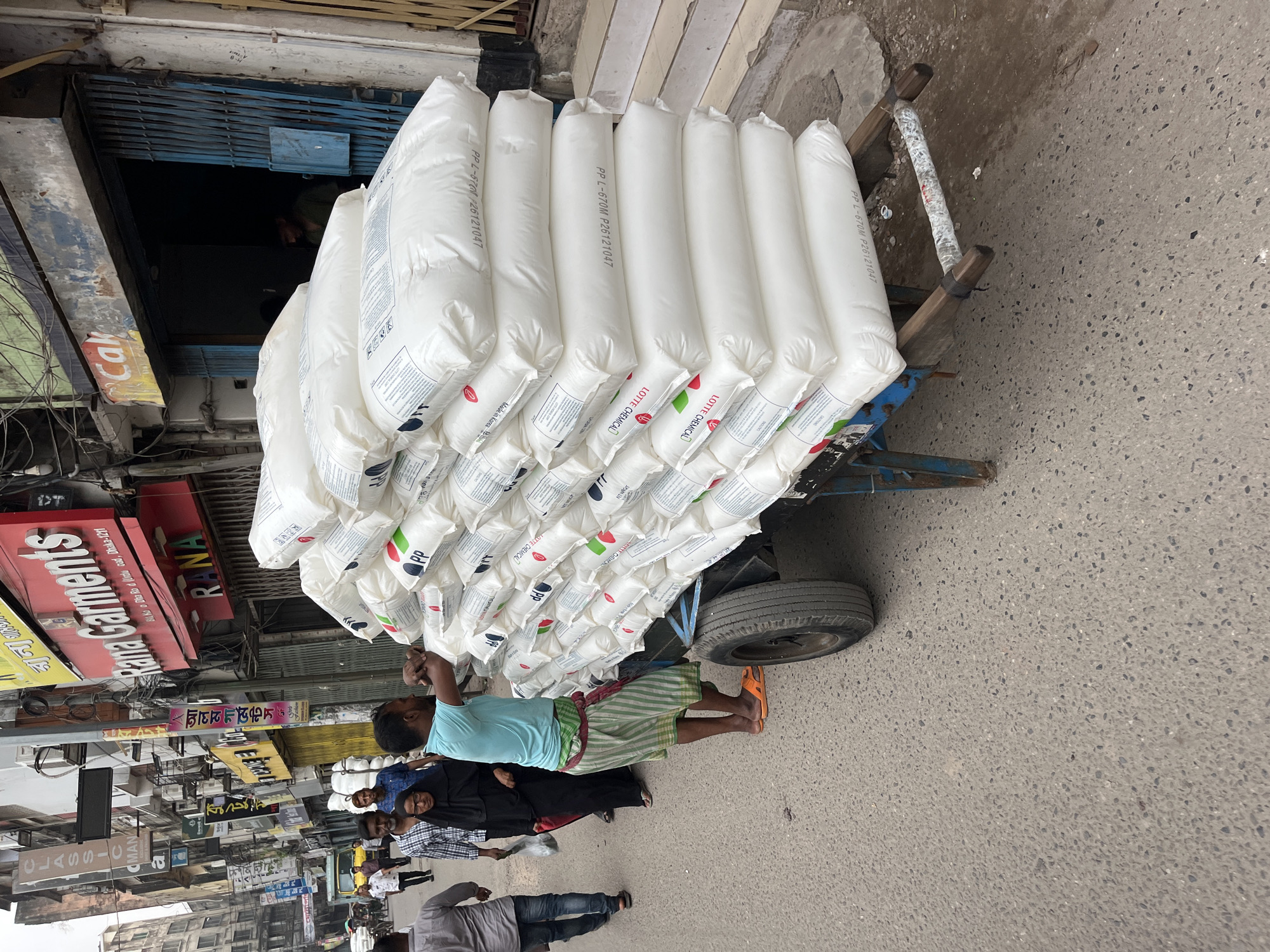 The image shows a street scene in Dhaka, Bangladesh. A man wearing a light blue shirt and striped lungi is standing on the street, apparently pulling a blue cart loaded with numerous large, white bags. The bags are labeled with text including "PP L-670M P26121047," "LOTTE CHEMICAL," and "Made in Korea." A woman in black attire and several other people are walking nearby. The street is lined with buildings featuring numerous signs, including "Rana Garments" and "CLASSIC MAN." There is a partly lowered shutter door to a shop. The street surface is paved.