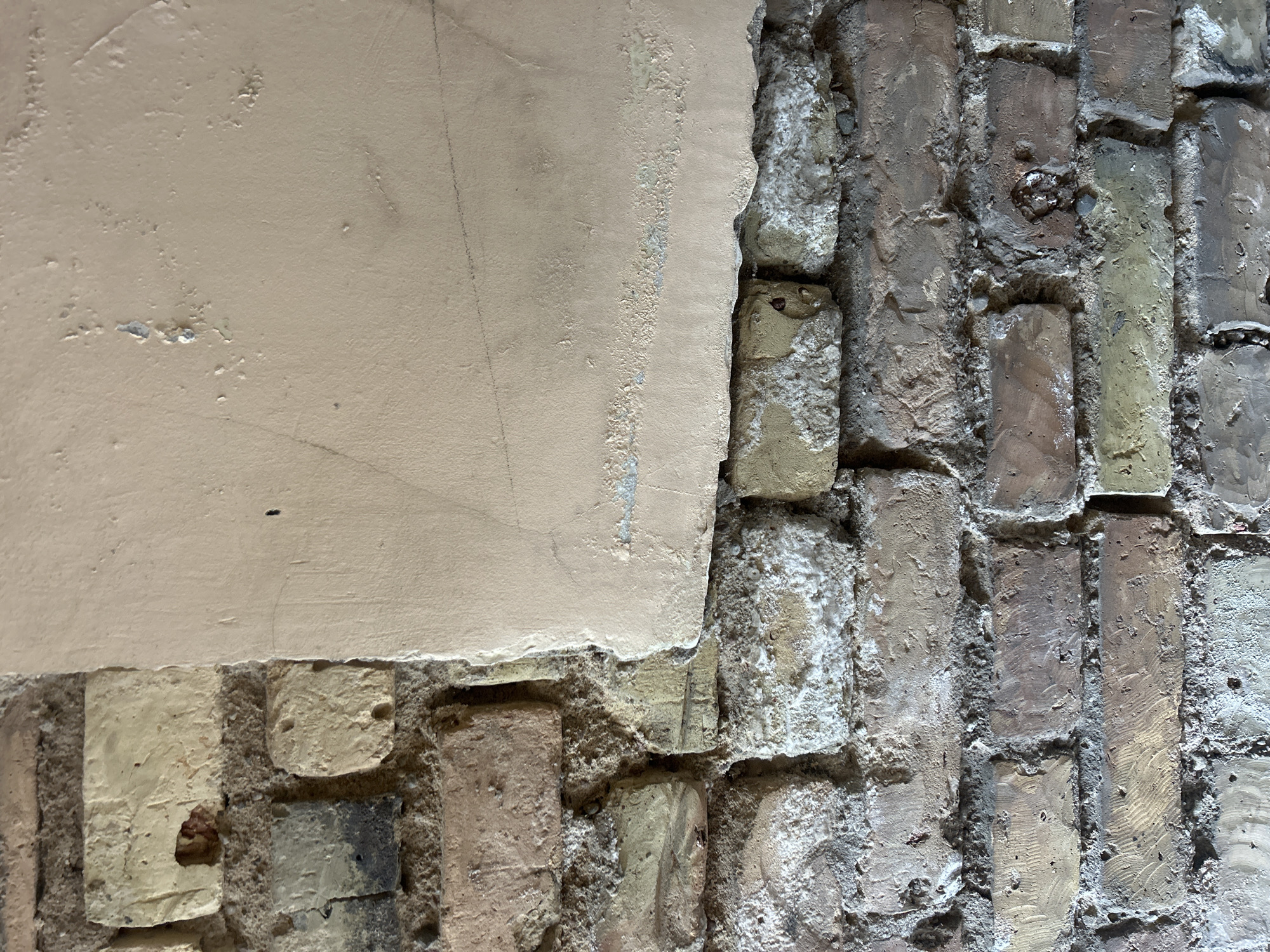 The image shows a close-up of a weathered brick wall partially covered by a rough, beige plaster. The bricks are varied in color, ranging from light yellow and beige to darker brown and gray, with some exhibiting signs of wear and damage. The plaster is peeling and chipped in places, revealing the brickwork underneath. There are faint pencil lines visible on the plaster surface. The scene appears to be an exterior wall of a building, possibly in an older part of a city. No people or specific actions are depicted. The image is framed vertically.