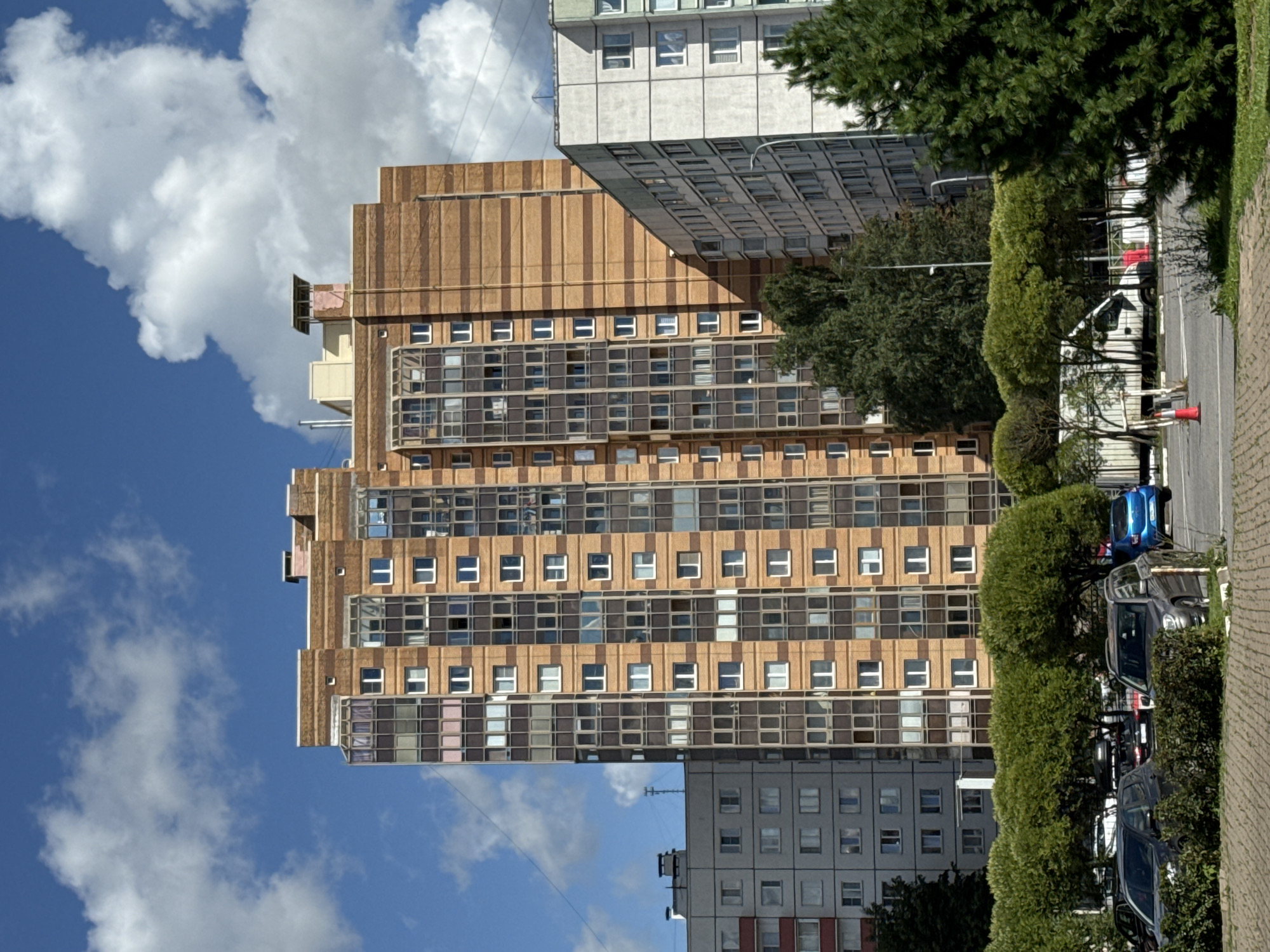 This is a medium-shot, eye-level, outdoor photograph of a residential building complex in Sinyavino, Russia, under a blue sky with scattered white clouds. The central focus is a tall, multi-story apartment building with a brick facade featuring horizontal banding of alternating shades of brown. The building has numerous windows, many of which are large and framed in dark material, creating a grid-like pattern. To the left of this building is a lighter-colored, multi-story apartment building with a simpler facade and smaller, more uniformly spaced windows. To the right, another apartment building with a light-colored facade and a more complex window arrangement is partially visible.
In the foreground, a row of meticulously trimmed green trees and bushes lines a parking area where several cars are parked. A dark gray SUV is prominent in the right foreground, and a bright blue car is visible in the background. Further back, a white truck with a metal cargo area is parked behind a fence. The ground in the immediate foreground is paved with textured concrete blocks. A few traffic cones are visible on the asphalt road in the middle ground. The overall impression is of a well-maintained urban residential area.