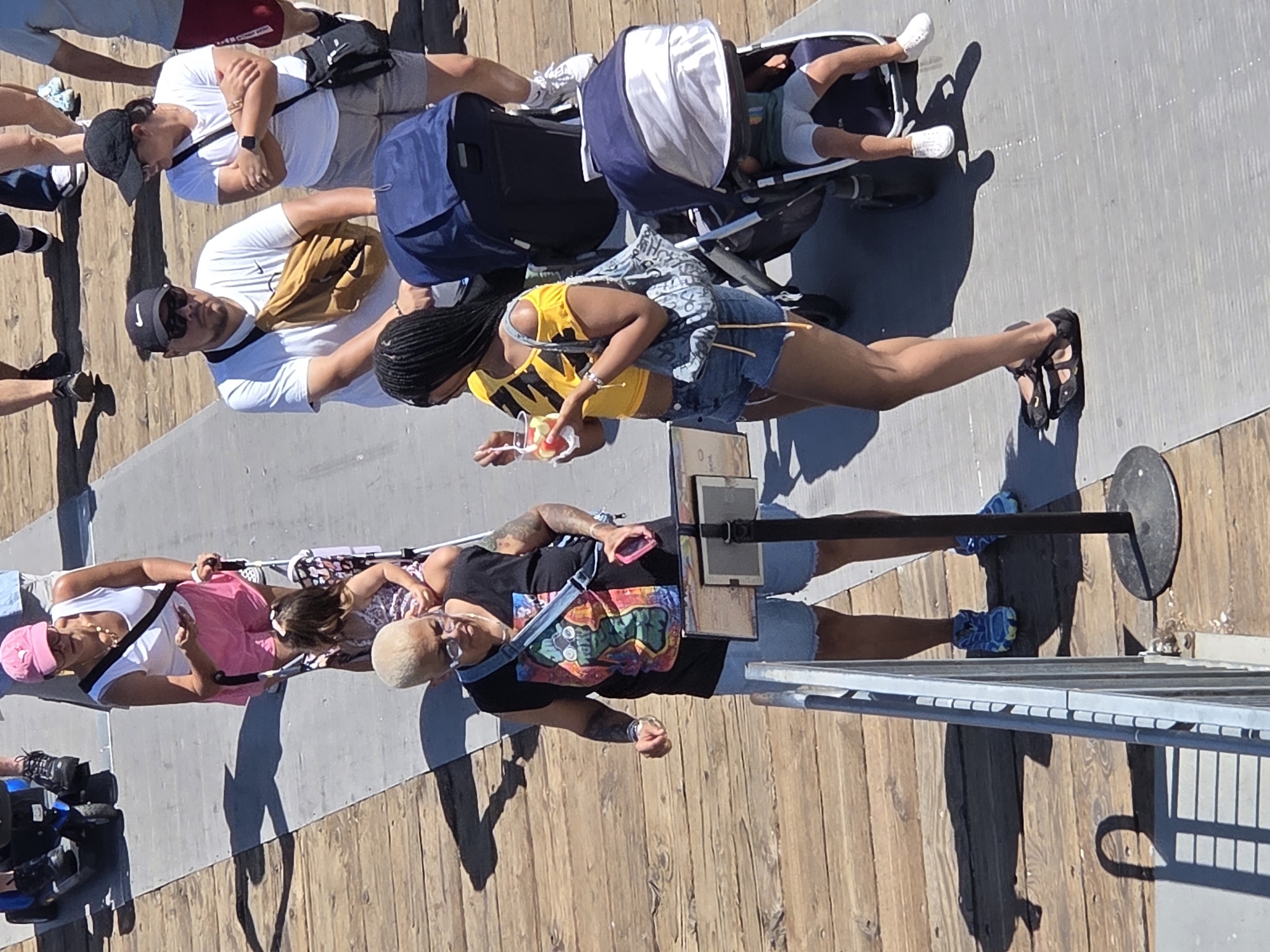 The image captures a scene on a wooden pier, likely in Santa Monica, United States of America, under bright daylight. Several people are walking or standing, with a few pushing strollers.
In the foreground, a person with short blonde hair and glasses is wearing a black sleeveless t-shirt with a colorful graphic print and denim shorts. They are holding a pink smartphone in their right hand and have a bag slung across their body. To their left, another person is walking, partially obscured, wearing a white tank top, pink shorts, and a pink baseball cap. They are pushing a stroller with a young child inside.
Further back, a person wearing a white t-shirt with a Nike logo, sunglasses, and a tan fanny pack is seen from the chest up. They are looking towards the left. Next to them, a person in a white t-shirt and a black baseball cap is looking at the first person and has their arms crossed. They are also wearing a black crossbody bag.
To the right, a person with long braided dark hair is wearing a yellow off-the-shoulder top and denim shorts. They are holding a cup with fruit and a spoon and have a patterned bag over their shoulder. They are standing next to a large dark blue stroller. Partially visible inside this stroller is a young child wearing a grey onesie and white shoes. Another stroller, a dark blue one with a grey canopy, is also present.
The ground is a combination of wooden planks and a grey paved surface. Shadows are cast by the people and objects, indicating the time of day is likely late morning or early afternoon. A metal railing runs along the left side of the image, and a black pole with a sign is positioned between the two people in the foreground.