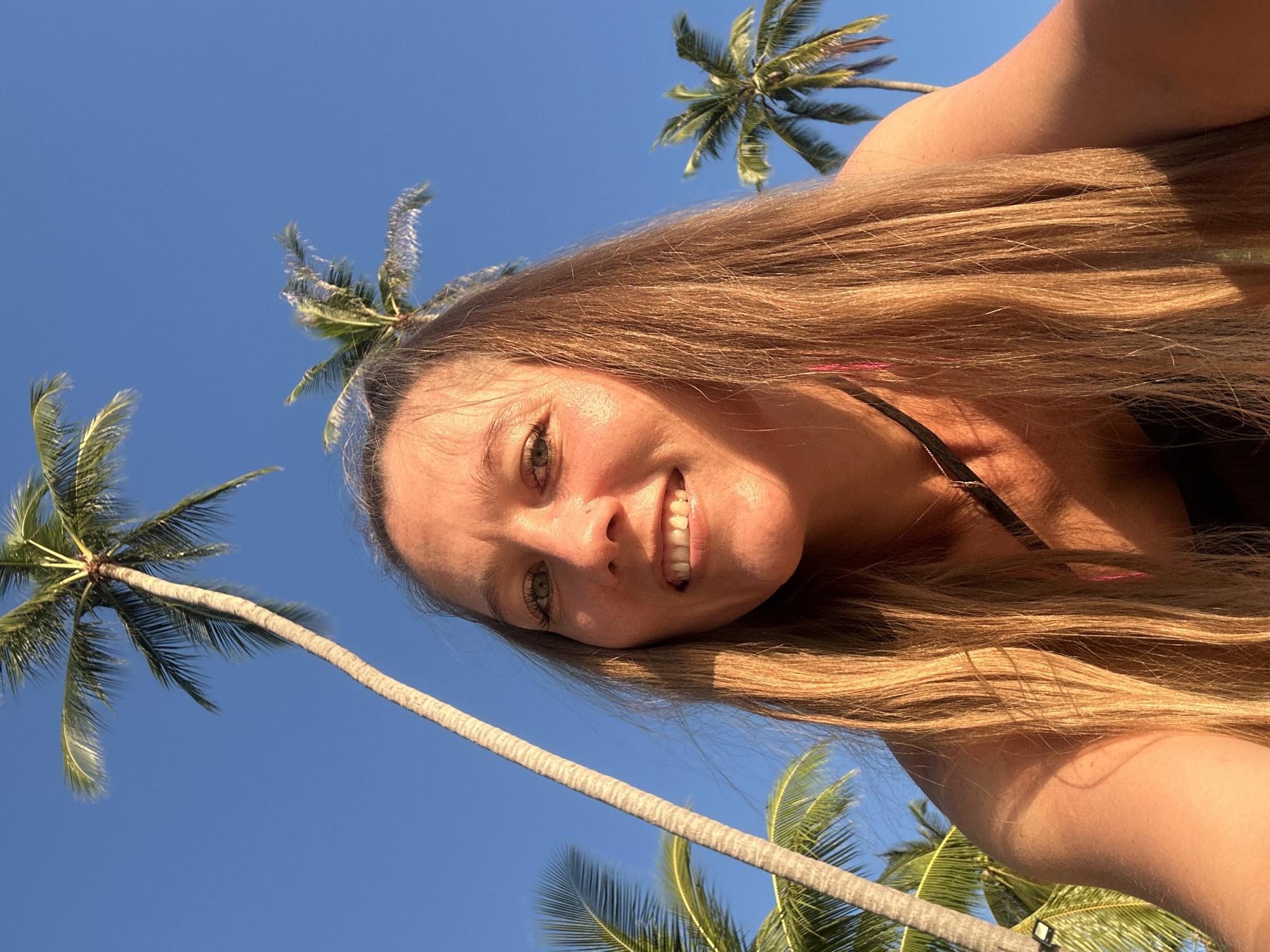 A woman with long, blonde hair smiles for a selfie outdoors under a clear blue sky, with several tall palm trees visible in the background. The setting appears to be a tropical location, possibly Baan Taling Ngam, Thailand, given the presence of palm trees and bright sunlight. The woman is looking directly at the camera, and her face is illuminated by the sun. She is wearing a black top with a thin strap visible.