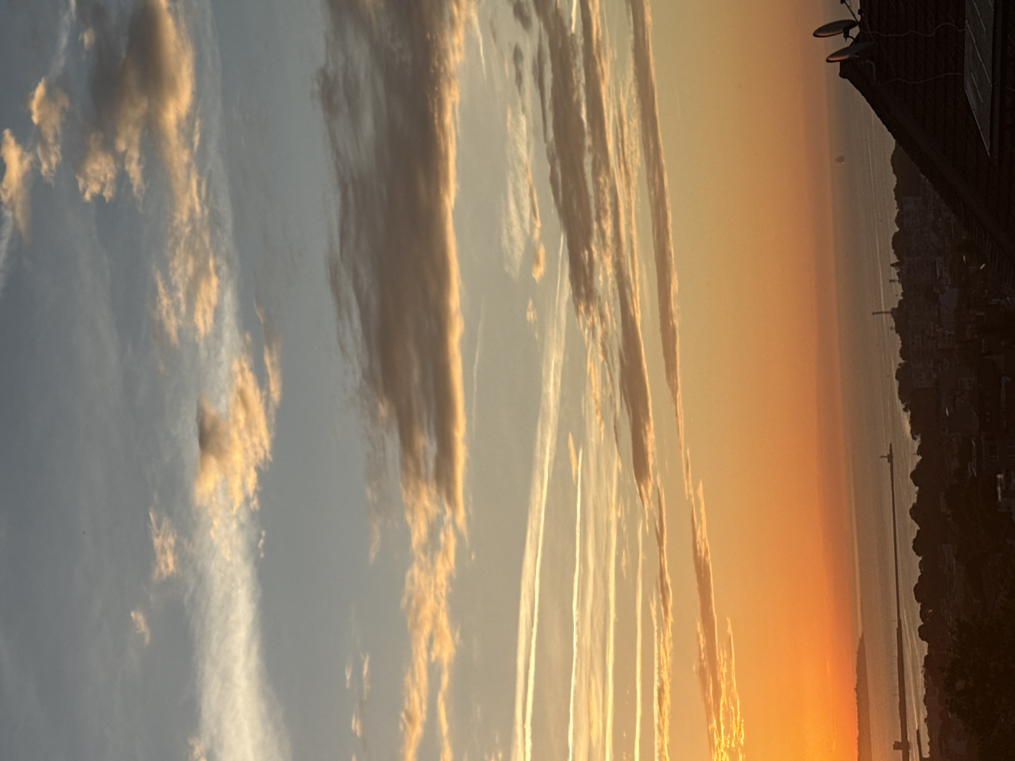 This is an aerial, vertical, full shot of a vibrant sunset over a coastal city, likely during the day. The sky is a gradient of warm oranges near the horizon, transitioning into soft blues and grays higher up. Wispy clouds, tinged with yellow and orange, are scattered across the sky. In the foreground, silhouetted against the bright sky, is a dark rooftop with two satellite dishes. Below the rooftop, the city is visible as a dense collection of dark buildings and trees. Beyond the city, a body of water, likely the ocean, stretches to the horizon. Several structures, possibly piers or breakwaters, extend into the water, and a lighthouse is visible on the right. A distant ship is also discernible on the water. The overall impression is one of a serene, picturesque coastal sunset.