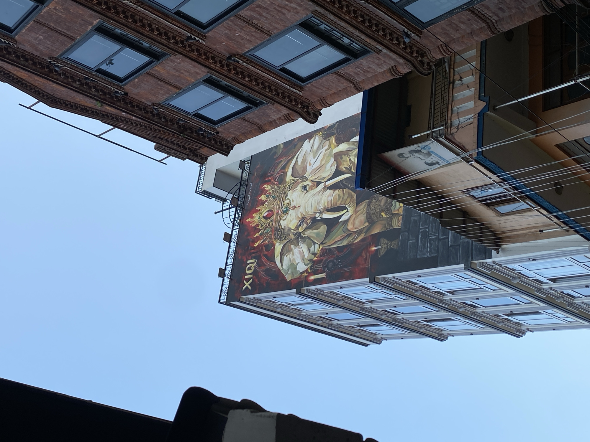 This image captures a low-angle perspective of an urban street scene in Kathmandu Metropolitan City, Nepal. Dominating the frame is a tall, multi-story building adorned with a vibrant mural of an elephant wearing a crown and ornate jewelry. The elephant is depicted in a stylized, almost divine manner, with a red background and fiery accents. Above the mural, the word "XIQI" is visible in large, white lettering.
To the right of this building, a narrower, older-looking structure with red brickwork and ornate detailing rises, featuring multiple windows. The contrast between the modern mural and the traditional architecture is striking. In the foreground and to the left, parts of other buildings are visible, adding to the dense urban feel. Electrical wires are strung across the scene, a common characteristic of city infrastructure. The sky above is a clear, pale blue. No people are clearly visible in the image. The overall impression is one of a bustling, culturally rich city where art and everyday life intertwine on building facades.