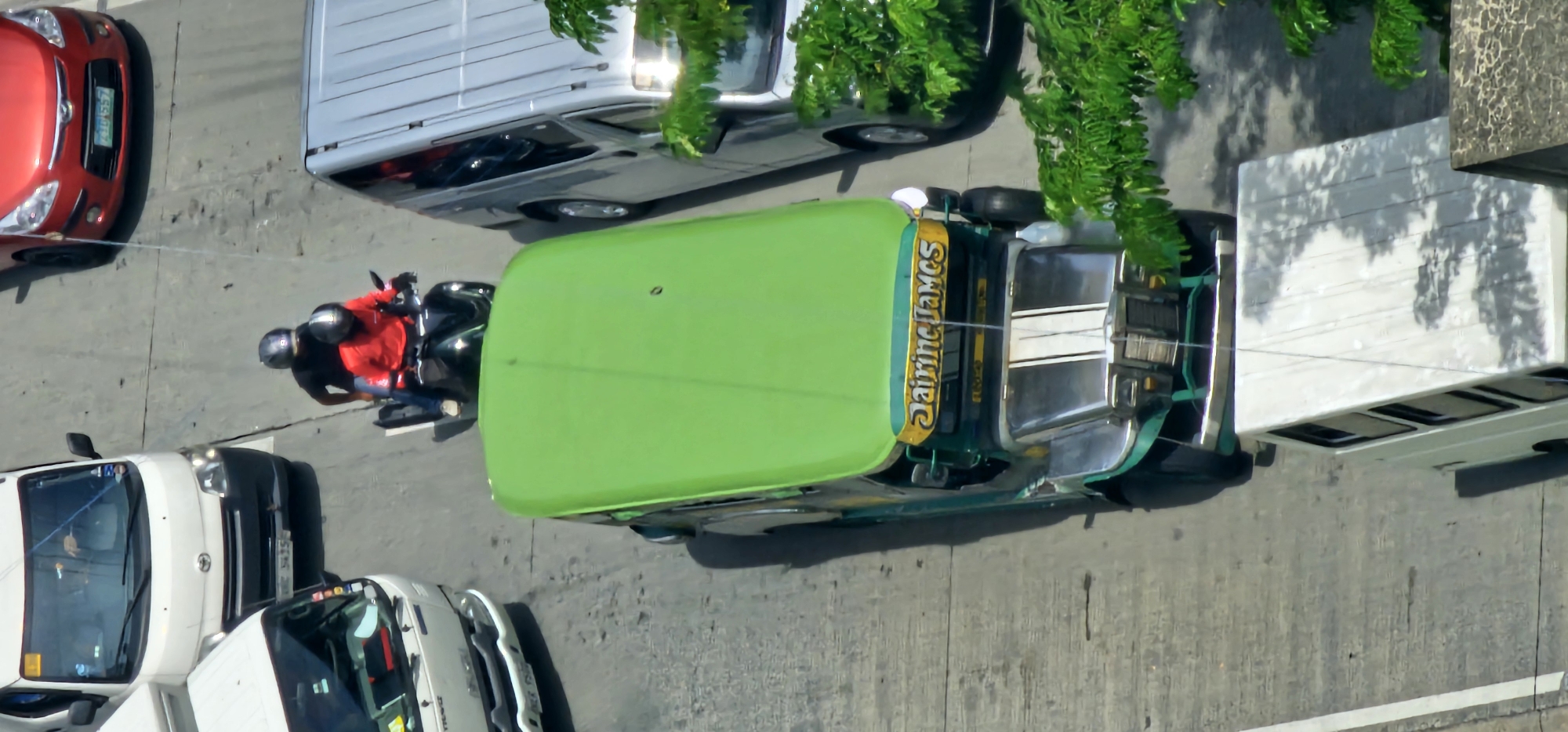An overhead, high-angle, vertical drone shot captures a traffic scene on an urban street in Makati, Philippines. In the foreground, a bright green jeepney, adorned with chrome accents and the name "Jairine James" in yellow lettering, occupies the center of the frame. To the right of the jeepney, a silver van is visible, its windows reflecting the surroundings. Behind the jeepney, a black motorcycle with two occupants wearing helmets is traveling. The rider in front wears a red shirt, and the passenger behind wears a black shirt. Further up the street, to the left, a white van is stopped. To the far right and slightly behind the red car, a segment of a red car is visible. The street is paved with concrete, showing wear and tear. Green foliage from trees encroaches on the right side of the image, casting shadows on the vehicles. The lighting suggests a bright, sunny day.