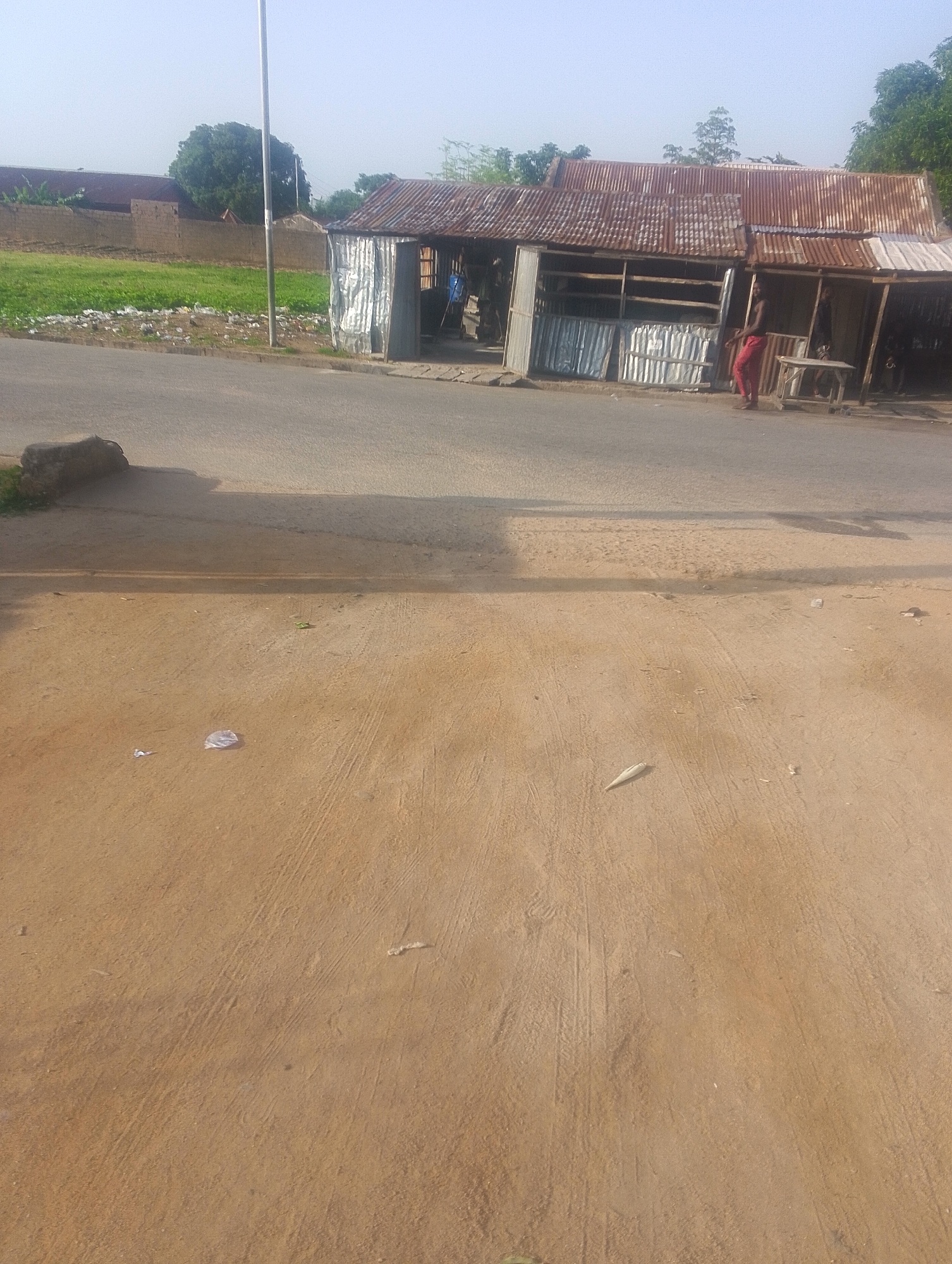 This is an outdoor, eye-level, full-shot photograph of a dusty road scene in Jalingo, Nigeria. The foreground is dominated by a tan, dirt ground with tire tracks visible. Some litter, including a crumpled plastic bag, is scattered on the ground. In the midground, a paved road runs horizontally across the frame, with a large, dark shadow cast from an unseen object to the left. A dark, rounded object, possibly a rock, rests on the edge of the road and the dirt.
In the background, a long, makeshift structure with a rusted, corrugated metal roof is situated along the side of the road. This structure appears to be a series of stalls or shops. One stall has a corrugated metal front, while another is partially open with wooden shelving visible. A person wearing red trousers is standing in front of one of the open stalls, facing to the right. Another person is partially visible behind them. To the left of the stalls, a brick wall and some greenery are visible, suggesting a property boundary. Beyond the wall, buildings with reddish-brown roofs and trees can be seen. A tall, thin pole, possibly a lamppost, stands vertically between the road and the wall. The sky is a pale, hazy blue, indicating a bright day.