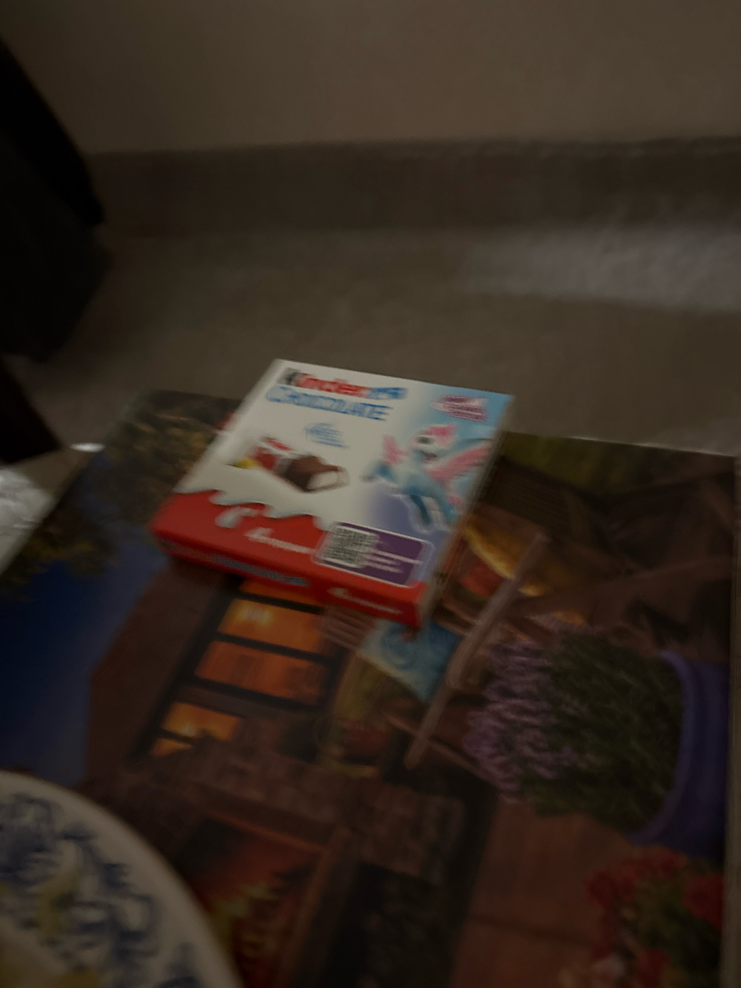 A blurry, overexposed, indoor, eye-level, medium shot shows a Kinder Chocolate box on a table. The box is red and white and features an image of a chocolate bar and a cartoon unicorn. The table surface is decorated with images of windows, flowers, and a blue pot. The background is dark and out of focus. There are no people or animals visible in the image. The location is not specified in Zavolzhye, Russia.