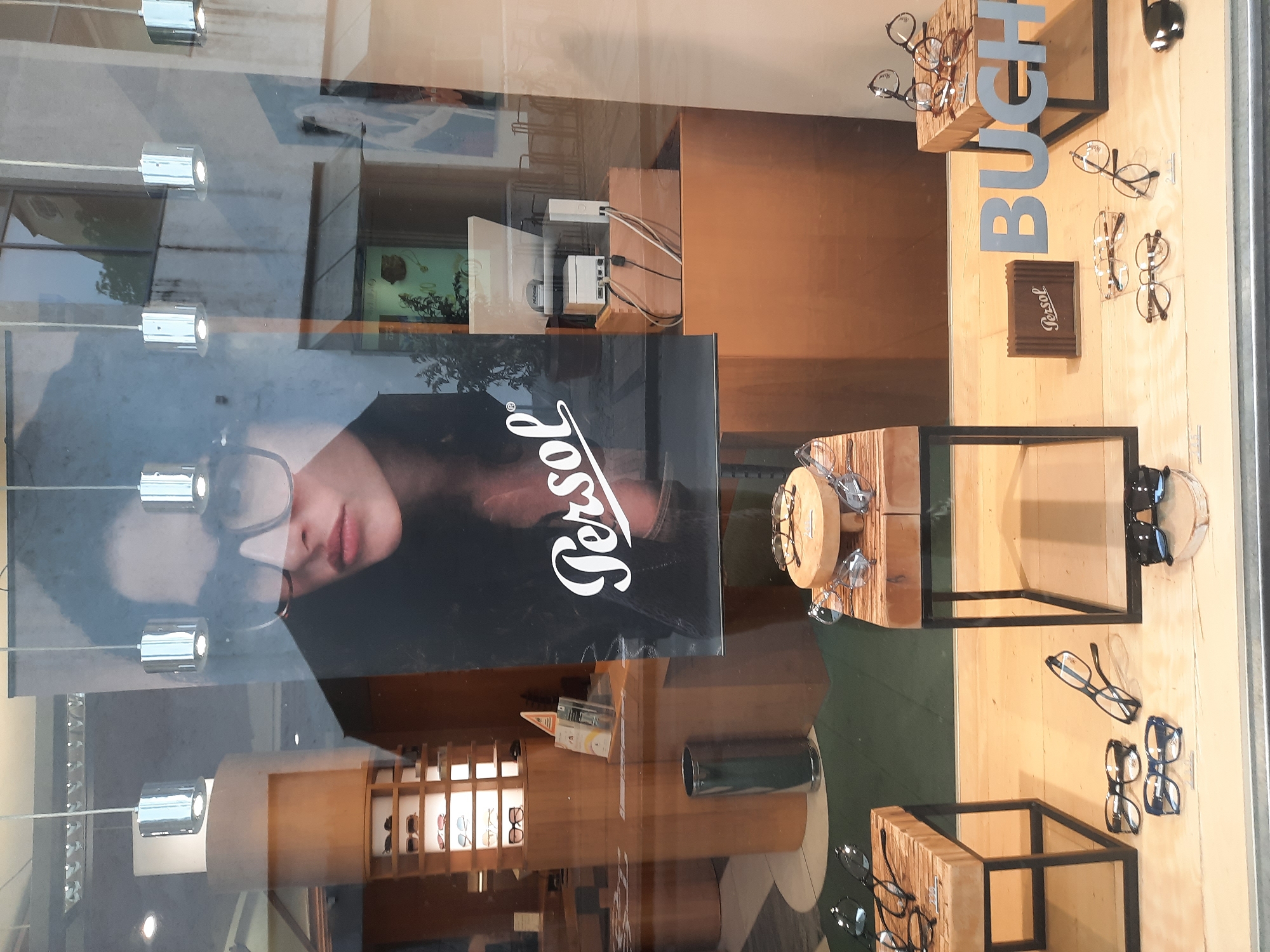 The image shows a shop window display of eyeglasses. In the background, there is a large advertisement featuring a woman wearing large framed glasses and the "Persol" logo. The shop interior visible through the window includes shelving units displaying more eyeglasses. In the foreground, there are several pairs of eyeglasses displayed on wooden stands and directly on the floor of the window. A wooden box with the "Persol" logo is also visible. To the right of the display, the word "BUCH" is visible, likely part of the shop's name or a brand. The display appears to be in a store located in Rosenheim, Germany. The scene is well-lit, suggesting it is daytime. The reflections in the window indicate it is an outdoor view of the shop.