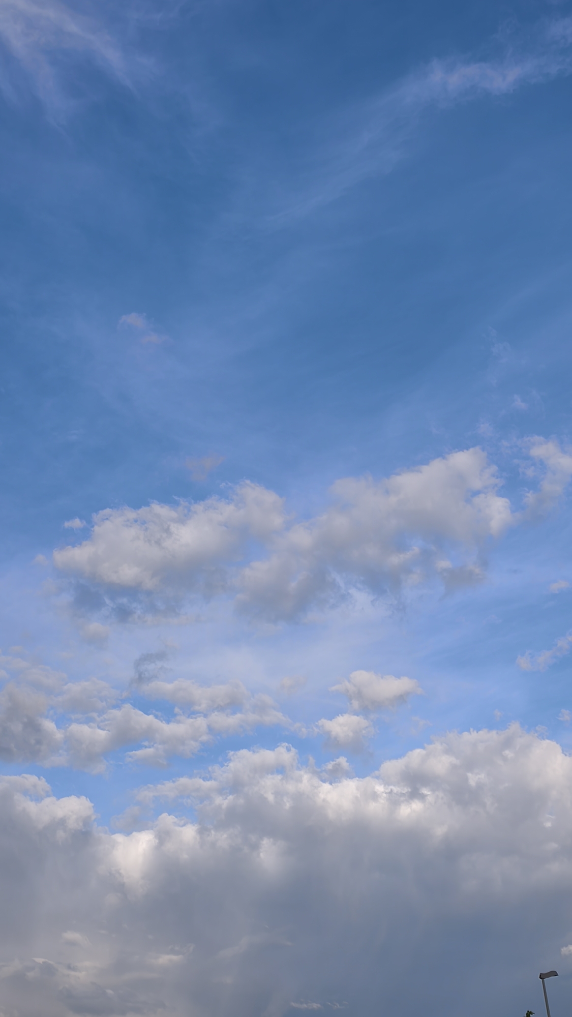 A vertically oriented image captures an expansive sky dominated by varying cloud formations over the city of Unknown, Unknown. The upper third of the frame features a clear, deep blue sky with sparse, wispy, translucent white cirrus clouds extending horizontally. The middle third transitions to a lighter blue, populated by scattered, distinct cumulus clouds, primarily white with subtle gray shading on their undersides, indicating depth. The lower third is filled with a dense, heavy layer of thick clouds, predominantly gray and white, with significant dark gray areas suggesting a lower, more opaque cloud ceiling or impending precipitation. In the bottom right corner, a segment of a dark gray or black streetlamp pole and a portion of its horizontal arm are visible. There are no identifiable people, animals, or other man-made structures present within the frame beyond the single streetlamp fragment. The scene is illuminated by natural daylight.