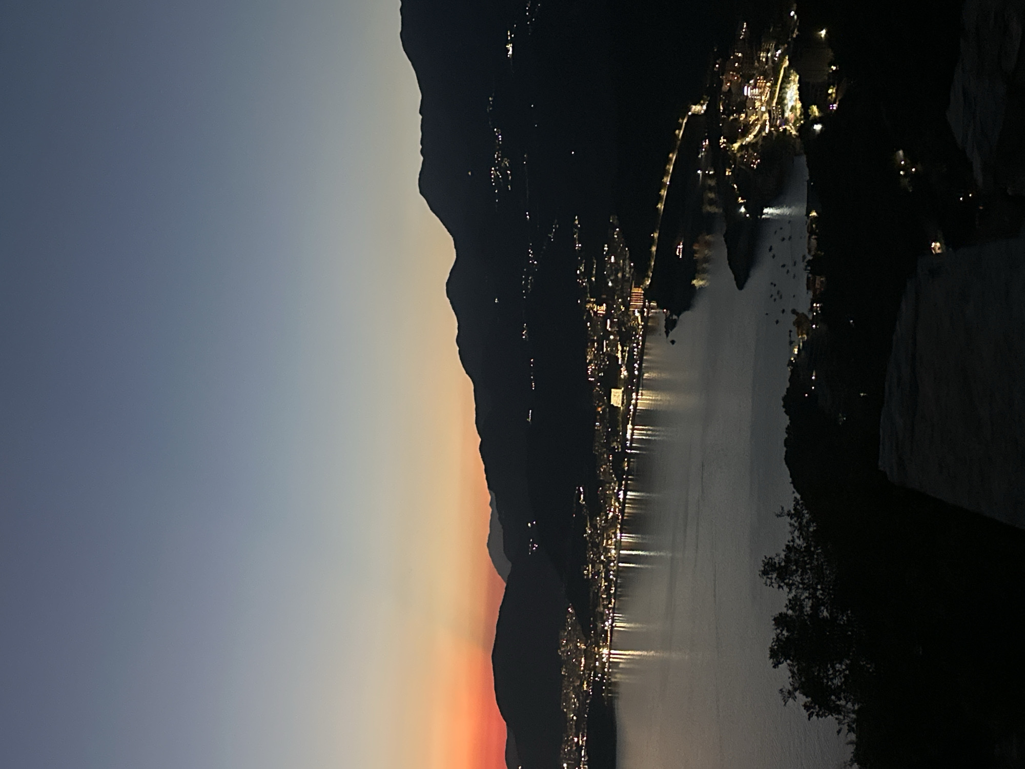 The image captures a twilight view of Sveti Stefan, Montenegro, from a high vantage point. The sky displays a gradient of soft blues and oranges, transitioning from the fading daylight. In the foreground, silhouetted trees and a rough stone surface are visible, suggesting a viewpoint on a hillside or rocky outcrop.
Below, the coastal town of Sveti Stefan is illuminated by numerous lights. Buildings and streetlights form a bright, continuous line along the coastline and extend onto the peninsula. The water, a calm expanse of dark grey, reflects the town's lights, creating shimmering patterns. Boats are dotted on the water, some appearing to be anchored.
Behind the town, a series of dark, mountainous silhouettes form the backdrop. Small clusters of lights are scattered across the slopes of these mountains, indicating human habitation or infrastructure. The overall scene conveys a sense of serene beauty as day transitions into night over the picturesque coastal landscape of Sveti Stefan.