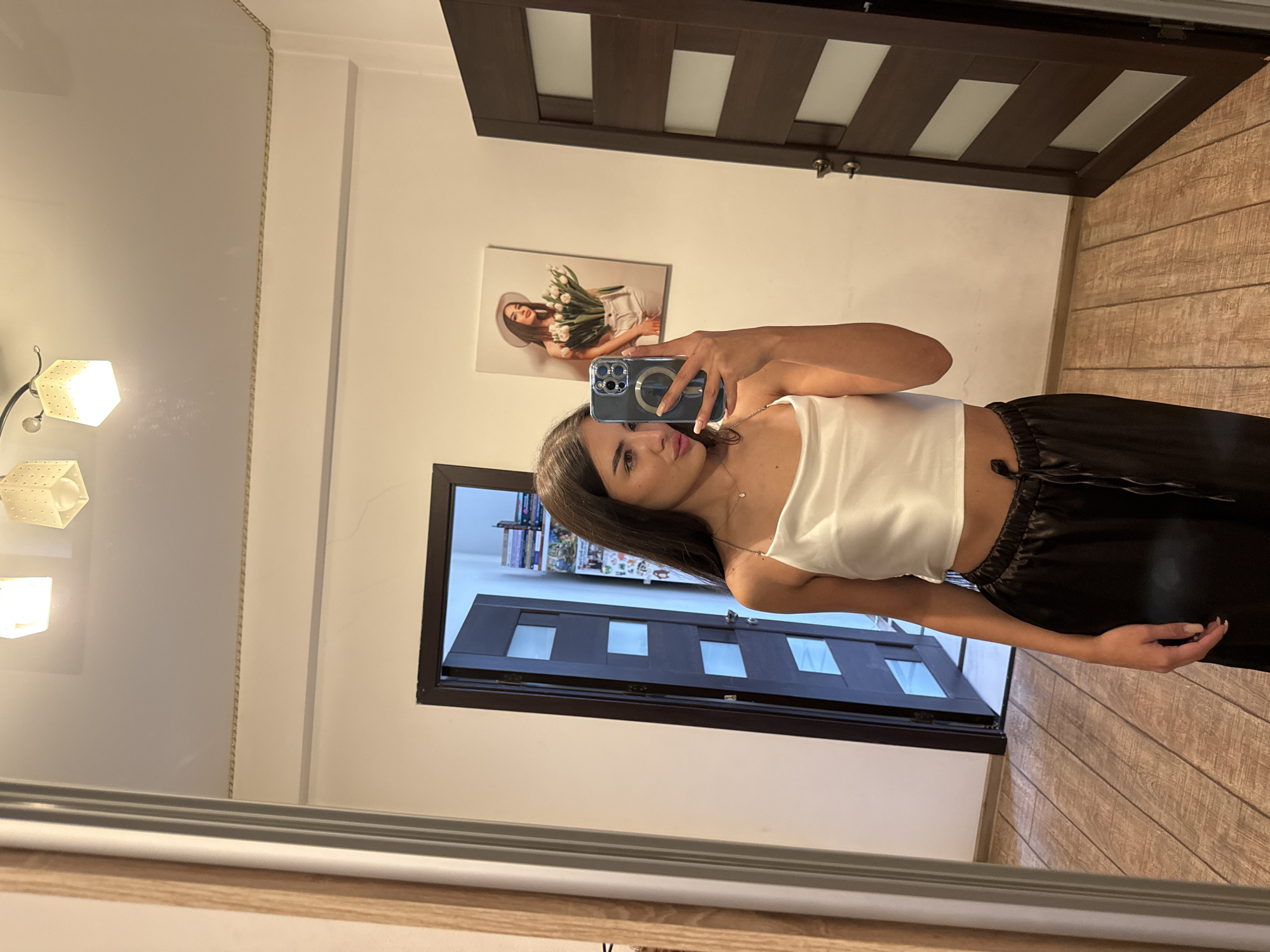 A young woman is taking a mirror selfie in a well-lit room. She is wearing a white satin crop top and black wide-leg pants. She is holding a smartphone in her right hand, angled to capture her reflection. In the background, a wooden door with frosted glass panels is slightly ajar, revealing a glimpse of a room with shelves filled with books. Another wooden door is visible on the right side of the frame. On the wall behind her, there is a framed picture of a woman holding a bouquet of white tulips. The room has wooden flooring and a decorative border along the ceiling. Two cube-shaped lamps are visible on the ceiling. The image appears to have been taken in an apartment in Chișinău, Moldova.