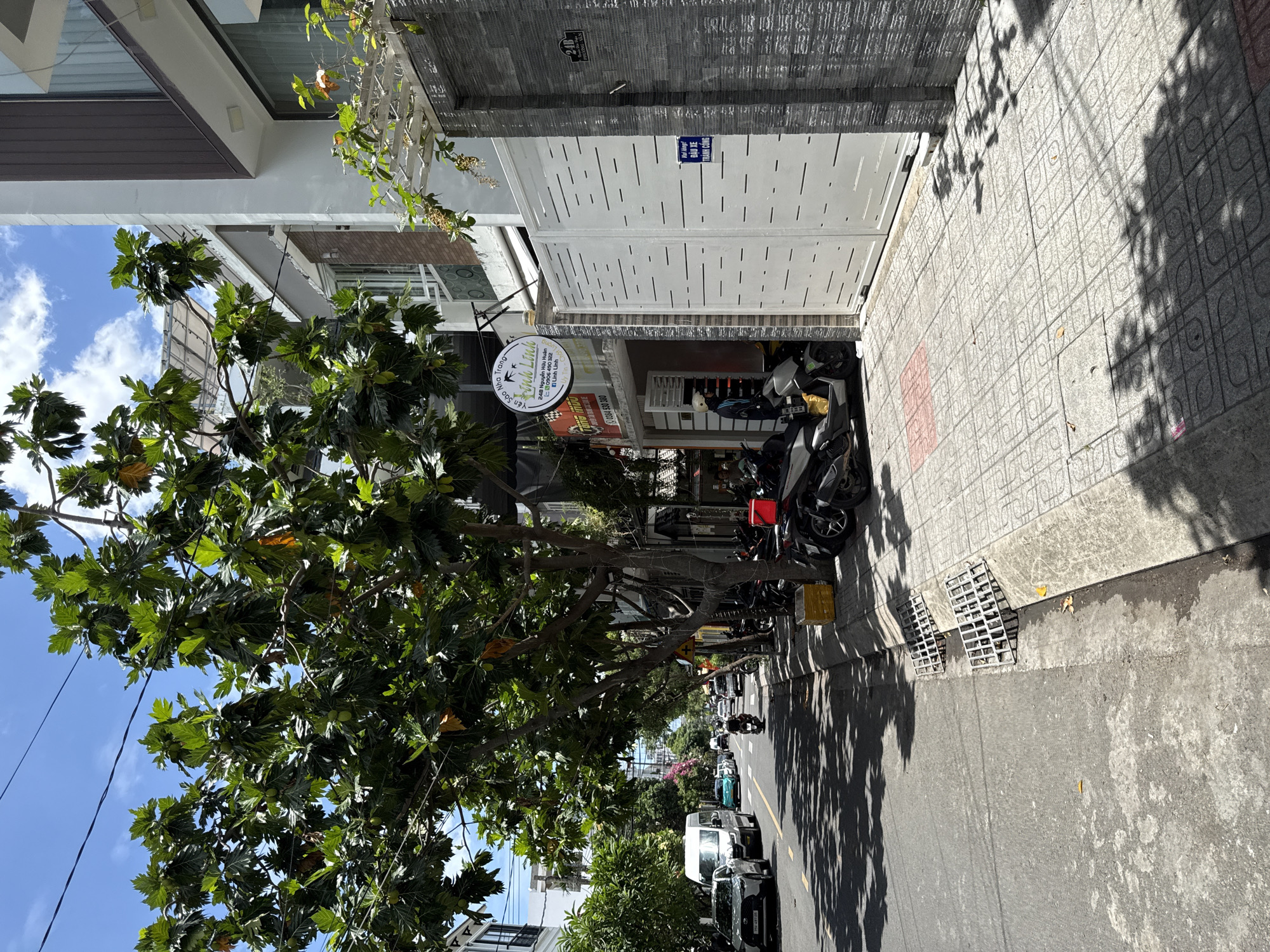 The image depicts a street scene in Nha Trang, Vietnam, on a sunny day. A large tree with green leaves and some brown, dried leaves shades a portion of the road and sidewalk. The street is paved with asphalt and marked with a yellow line. Several vehicles are visible on the road, including a white van, a silver car, a teal car, and a motorcycle with a rider. Parked along the sidewalk are several motorcycles.
To the right of the street, a sidewalk is paved with grey square tiles with a patterned design. In front of a modern building with white and grey stone cladding and a brown section on the upper floor, a large white gate with horizontal slits is visible. A small blue sign with Vietnamese text is attached to the gate. Adjacent to the gate, there is another sign with a circular logo and Vietnamese text that appears to indicate a business name. A person wearing a white helmet and a dark jacket is standing on the sidewalk next to a parked motorcycle.
The building to the right has large windows and a trellis structure with climbing plants above it. The environment suggests an urban setting with a mix of residential and commercial properties. The lighting indicates it is daytime, with distinct shadows cast by the trees. The sky is blue with scattered white clouds.