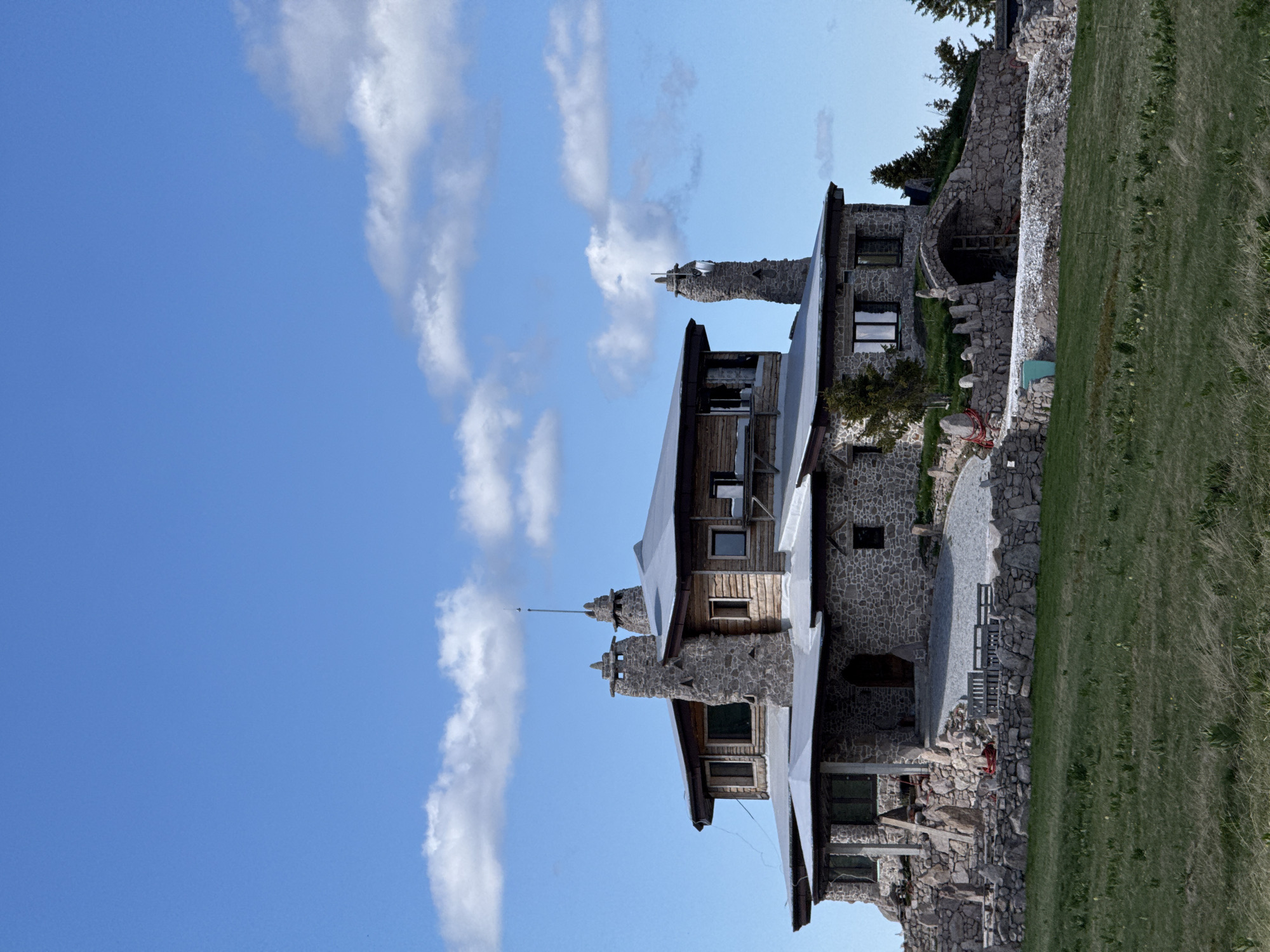 The image displays a large, multi-story stone and wood building with a complex roofline, situated on a grassy hill under a bright blue sky with scattered white clouds. The building features numerous stone chimneys, prominent stone foundations, and a mix of stone and wooden exterior walls. Large windows are visible on various levels. The surrounding environment includes more stone landscaping, a gravel path leading to an arched entrance, and some sparse vegetation, including trees on the right side. The foreground is dominated by a green, somewhat dry grassy field. There are no people or animals visible in the scene. This building is located in Gela, Bulgaria.