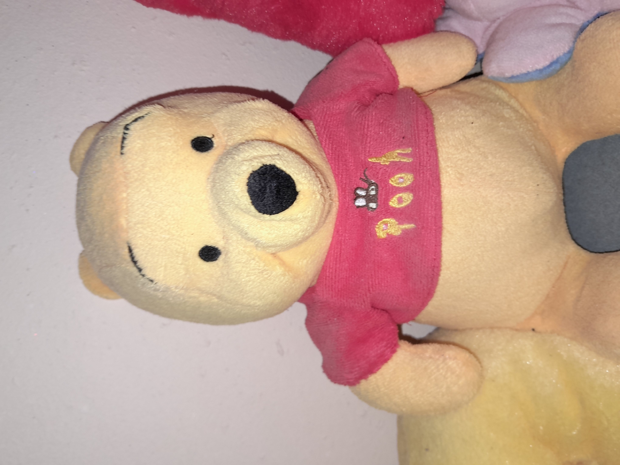 A stuffed Winnie the Pooh toy is positioned centrally in the frame, facing forward. The toy is yellow and wears a pink t-shirt with the word "Pooh" embroidered in yellow, along with a small bee graphic. Behind the Winnie the Pooh toy, to the right, a portion of a pink stuffed toy is visible. To the left, a larger, out-of-focus yellow object is present. The background is a plain, lightly textured light-colored wall. The image appears to be an indoor shot, likely in a domestic setting, with no visible indicators of the city of Uchquduq, Uzbekistan.