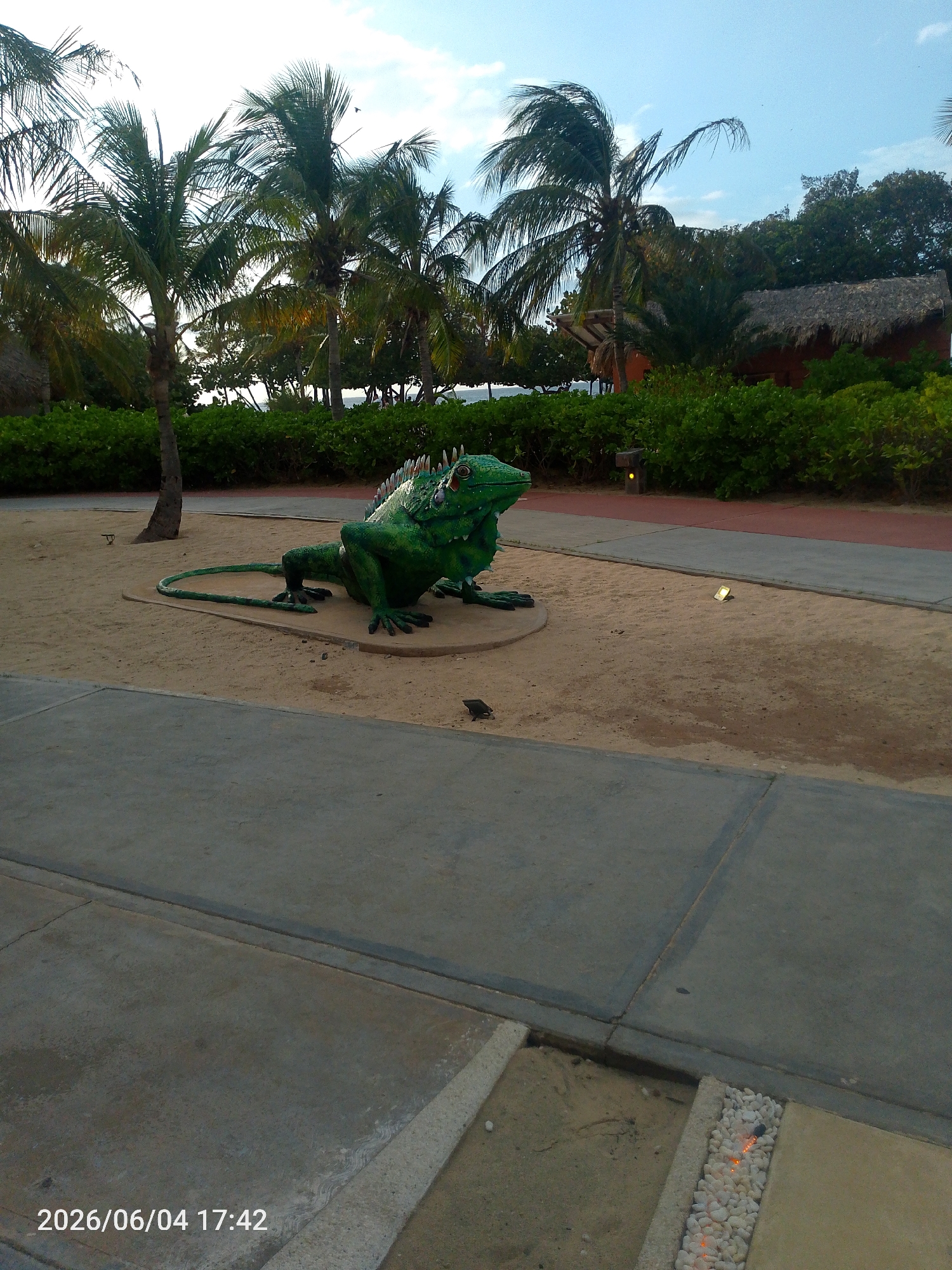 The image captures an outdoor scene, likely in a tropical or resort setting, during the late afternoon. The central subject is a large, vibrant green statue of an iguana or similar lizard, prominently displayed on a concrete base within a sandy area. The iguana statue features distinctive dorsal spines and is oriented towards the right side of the frame.
The ground consists of a combination of light grey concrete pathways and sandy patches. A wide concrete path dominates the foreground, while another runs horizontally across the midground, with a section of reddish-brown pathway visible further back. Several small, ground-level lights are embedded in the sand and along the paths, one emitting a distinct yellow glow. In the bottom right corner, a decorative strip of small white pebbles contains several warm-toned embedded lights. A small, dark object, possibly a bird or another miniature statue, is visible on the sand near the iguana's tail.
The background is lush with vegetation, including numerous tall palm trees with green fronds that stretch towards a bright, slightly cloudy sky. Dense green bushes form a hedge-like barrier in the midground, separating the immediate area from the distant elements. Partially obscured by the foliage on the right, a building with a dark, thatched roof adds to the tropical ambiance. A glimpse of a body of water, presumably the ocean or sea, can be seen beyond the greenery on the left side.
No people are present, and no specific activity or event is taking place. The scene conveys a sense of calm and a vacation-like atmosphere. A timestamp "2026/06/04 17:42" is visible in white text at the bottom left corner, indicating the time of day is 5:42 PM, which aligns with the gentle, diffused lighting.
