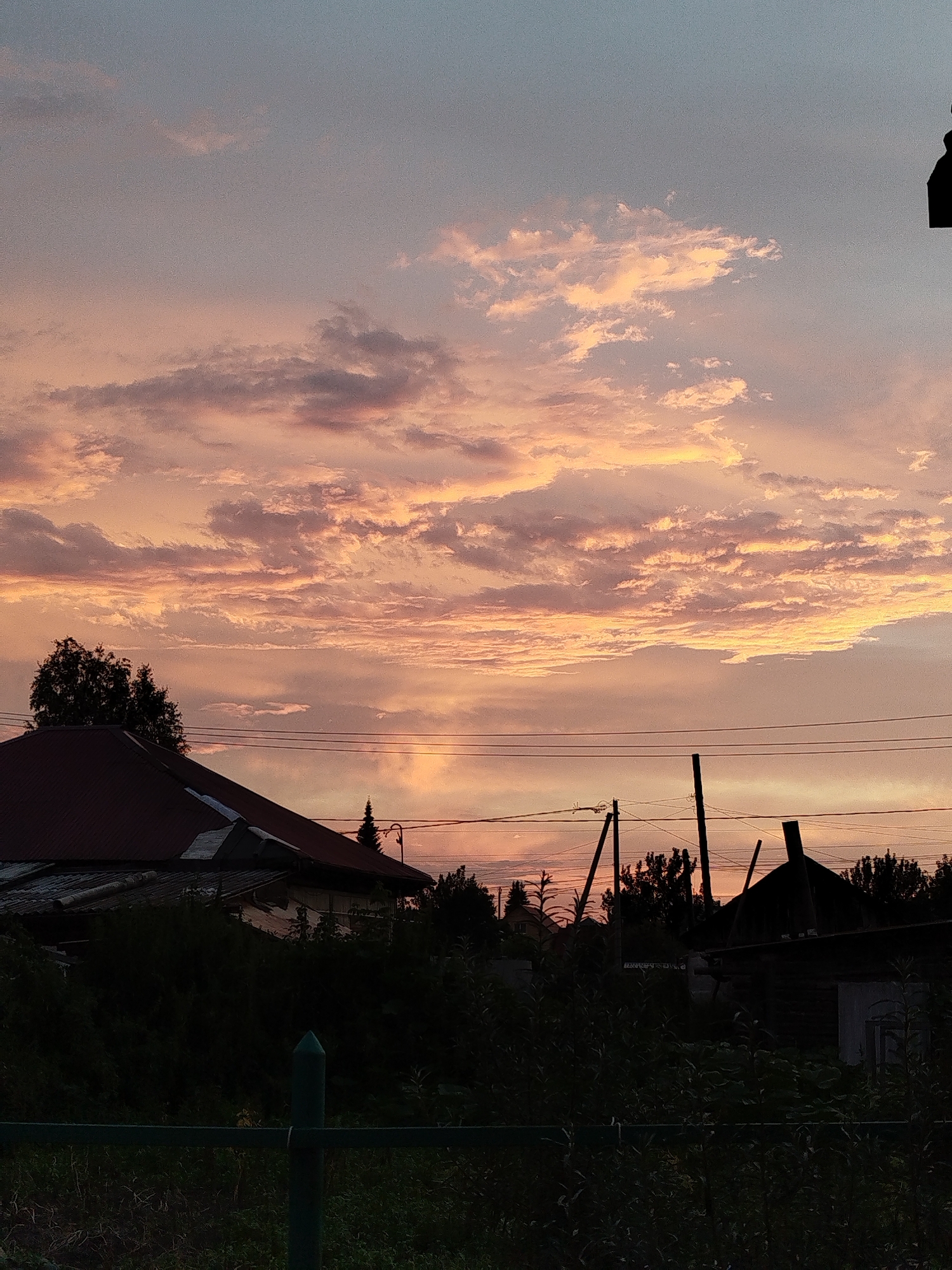 The image depicts a rural or suburban landscape at dusk. A sunset is visible in the sky, casting warm hues of pink, orange, and yellow through scattered clouds. Silhouetted against the sky are power lines, telephone poles, and the roofs of houses and other structures. In the foreground, dark foliage and a green fence partially obscure the view. The overall impression is one of a peaceful evening in a sparsely populated area, possibly on the outskirts of Zavodoukovsk, Russia, given the context.