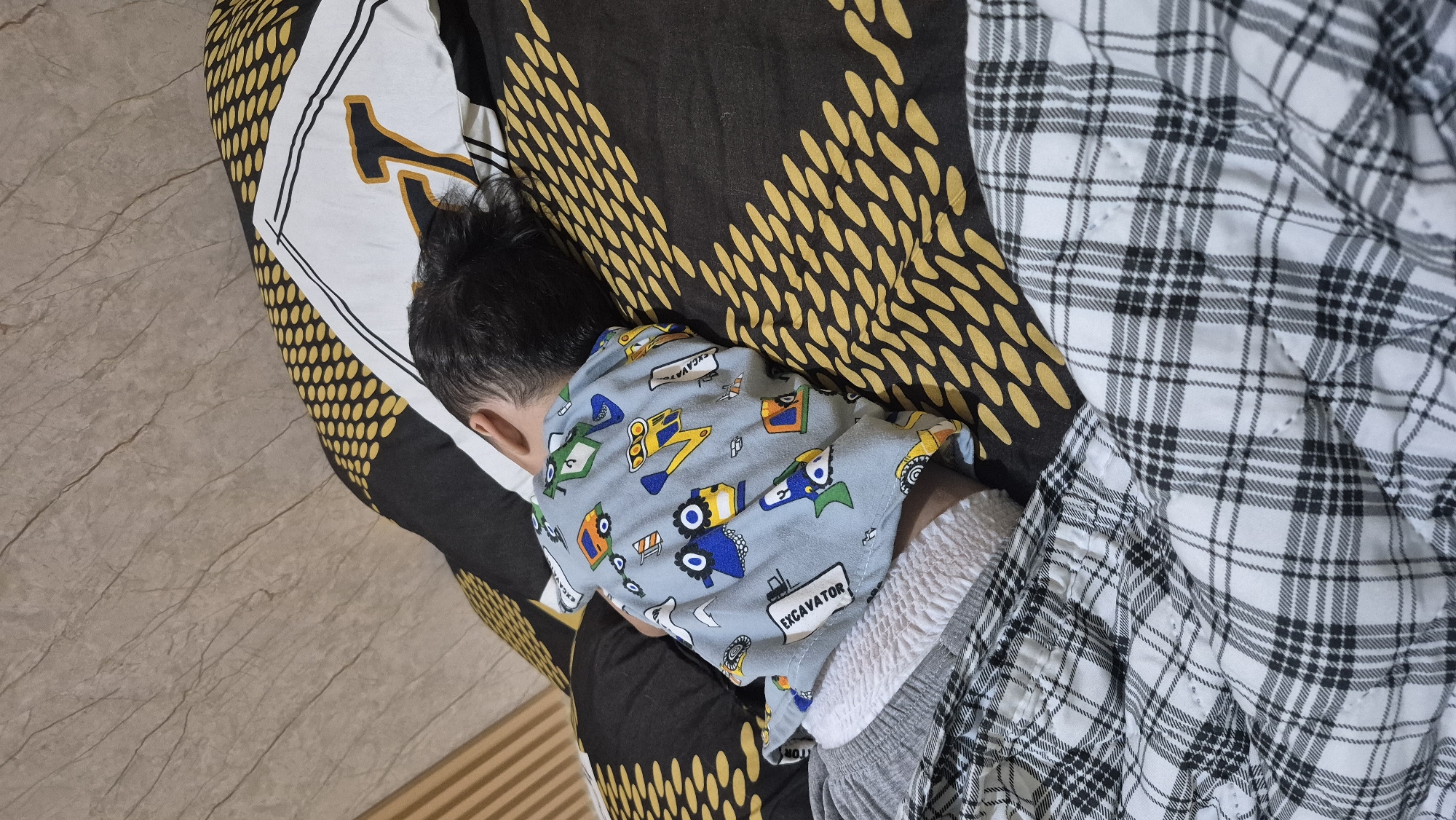 A young child is sleeping on a bed. The child is lying on their side, facing away from the camera, with their head resting on a pillow. They are wearing a light blue pajama top with a print of various construction vehicles, including excavators and trucks, and the word "EXCAVATOR" is visible on one of the prints. The child is also wearing gray pajama bottoms.
The bed is covered with patterned bedding. The pillowcase has a black and yellow abstract design with white borders. The blanket or duvet has a black and yellow pattern consisting of many oval shapes, and a section of black and white plaid fabric is visible on the right side of the image.
The background appears to be a floor with a marble-like pattern, in shades of beige and gray. A wooden slatted object is visible in the lower right corner, possibly part of the bed frame or furniture.
The scene suggests a quiet moment, likely during the daytime or evening, as the child is asleep. There are no strong indicators of the weather or time of day beyond the fact that the child is in pajamas and sleeping. The overall impression is one of comfort and rest. The location context is South Tangerang, Indonesia.