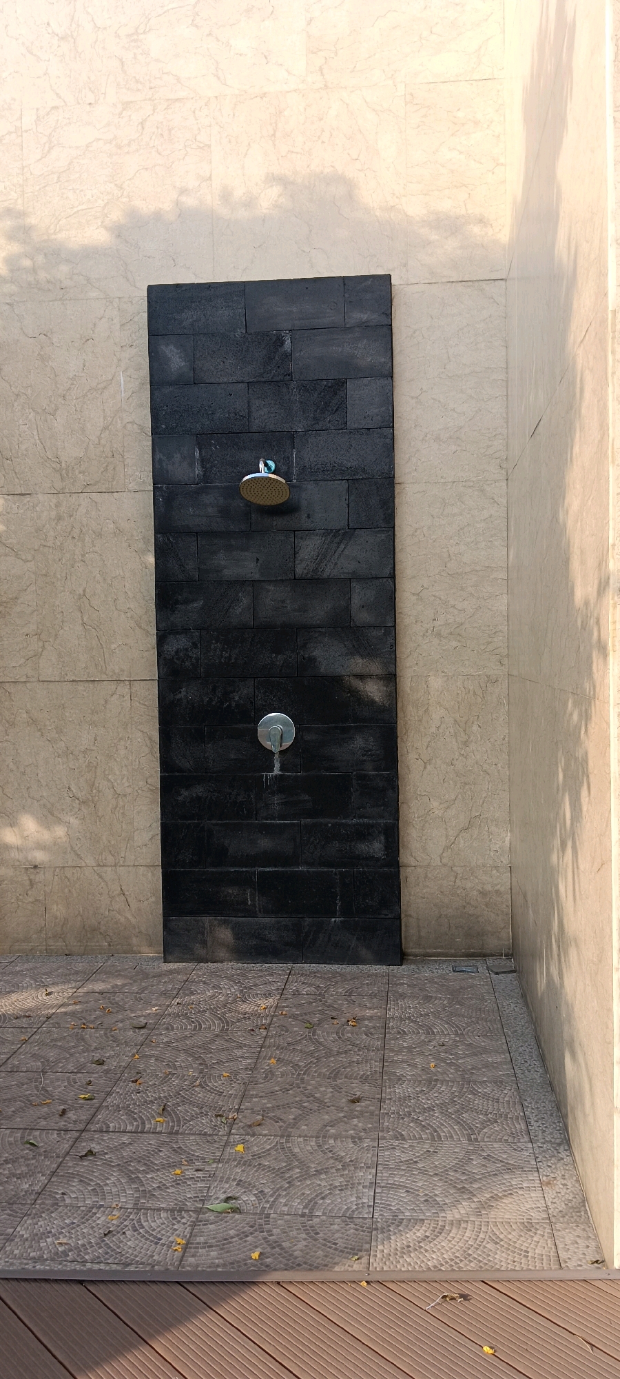 In Kemang, Indonesia, an outdoor shower area is depicted. The shower is set against a vertical panel of dark, stacked stone tiles, contrasting with the surrounding light beige, marble-patterned wall. A circular showerhead is positioned near the top of the dark panel, and a silver faucet with a single handle is located below it, with traces of water dripping. The ground in front of the shower is tiled with a mosaic pattern of grey and white circles. To the right, a section of light beige wall is visible, casting a shadow from what appears to be overhead foliage. In the foreground, there is a wooden deck. The scene is illuminated by natural light, creating dappled shadows on the walls and ground.