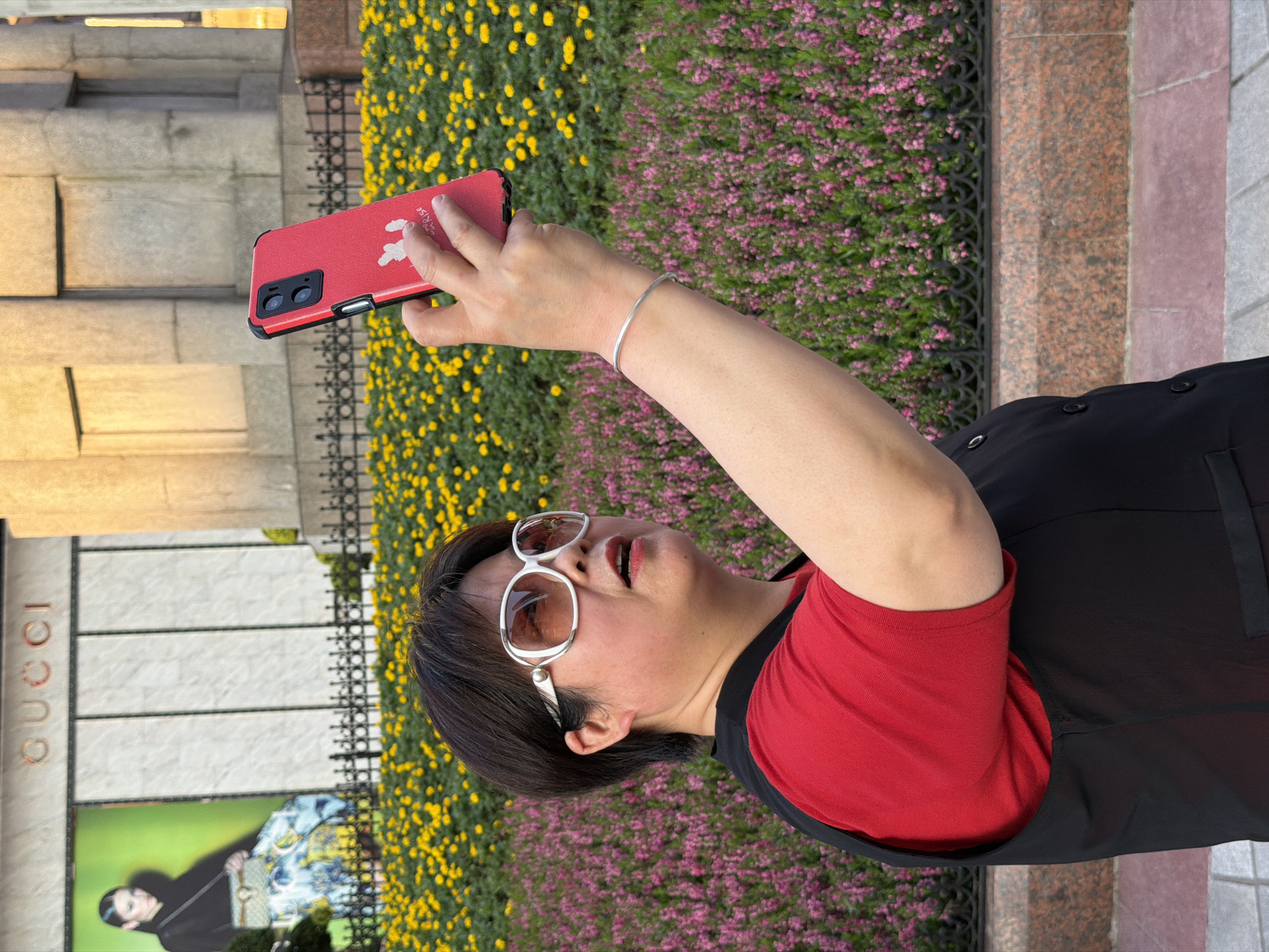 A woman with short, dark hair and large white-rimmed sunglasses is taking a selfie with her red smartphone in front of a Gucci store. She is wearing a red t-shirt and a black vest. Behind her, a billboard displays a model in a black top and a patterned skirt, with a green background. A black metal fence separates the foreground from a flower bed with yellow flowers and a lower flower bed with pink flowers. The building in the background appears to be made of stone. The scene is set in the city of Yuzhong District, China.